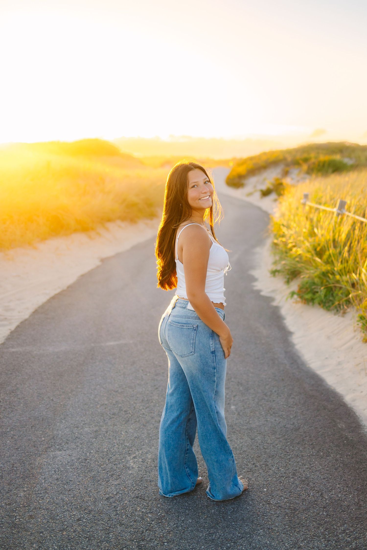 A high school senior in denim jeans walks along a coastal path during golden hour sunset for senior photos at Horseneck Beach in Westport MA
