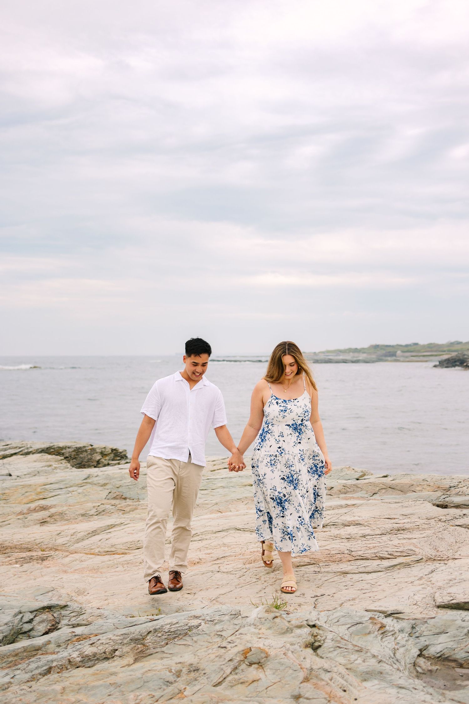 Newly engaged couple walking hand in hand along rocky coastal shore with ocean and cloudy sky on Ocean Drive in Newport Rhode Island