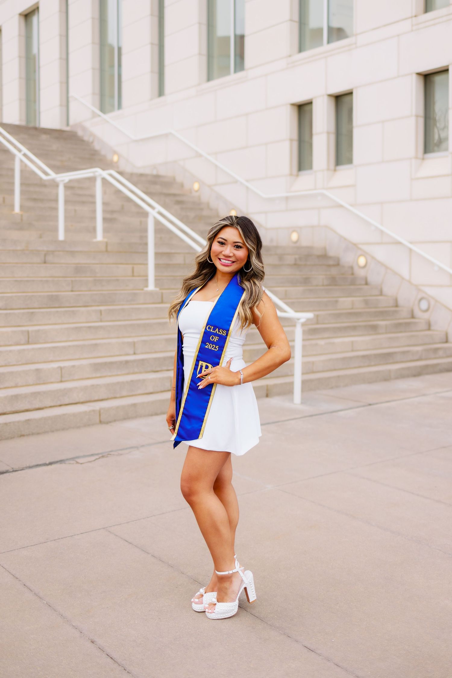 Graduate in white dress and blue sash poses on concrete stairs outside modern school building for grad photos at UMASS Boston Massachusetts