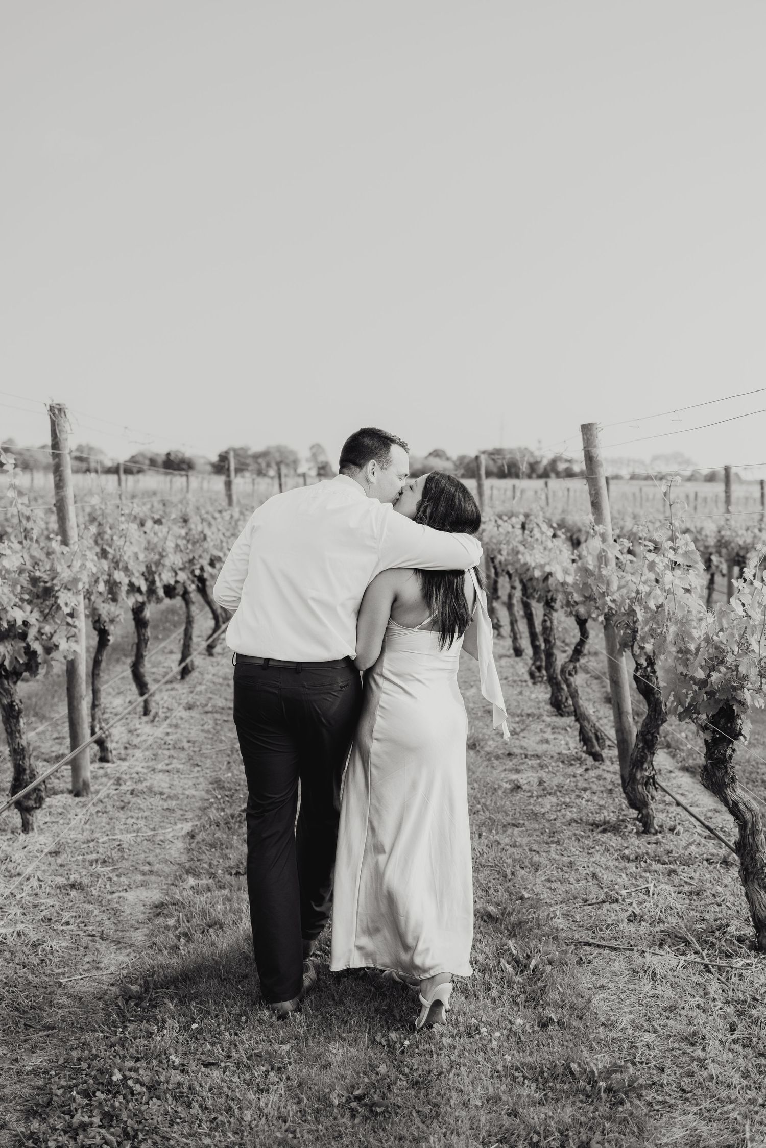 A couple embraces while walking through rows of grapevines in a vineyard in black and white photography.