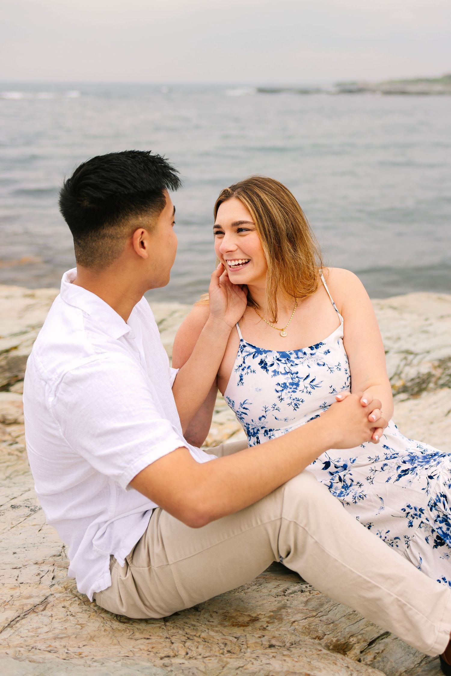 An engaged couple sits on coastal rocks by the sea on Ocean Drive in Newport RI, sharing a joyful moment after a surprise proposal.