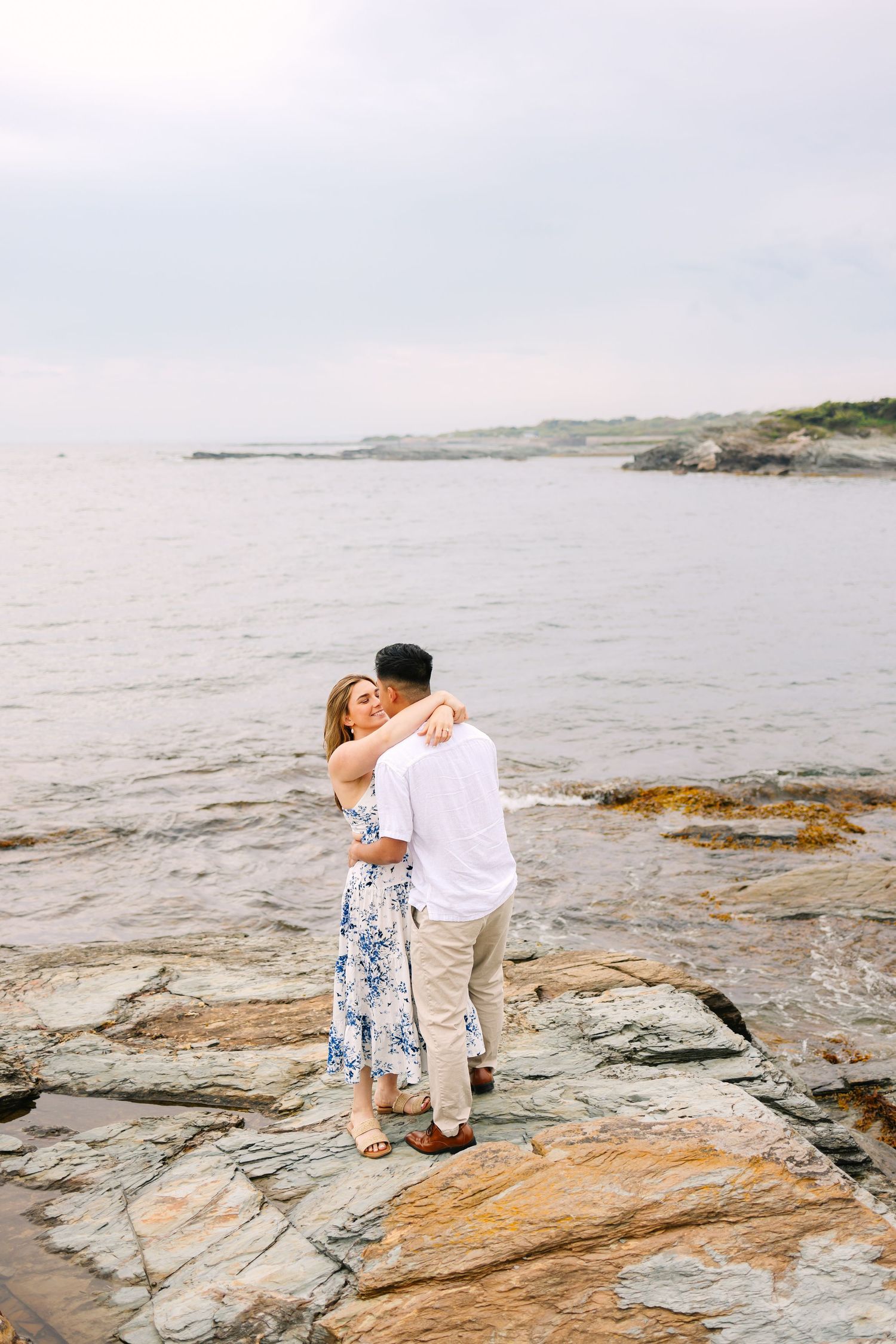 Couple embracing romantically on rocky coastal shoreline for engagement photos with ocean backdrop in Newport Rhode Island Ocean Drive