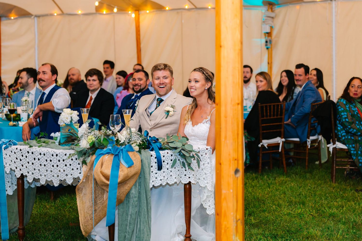 A couple sits together at their outdoor tent wedding reception with guests seated behind them.