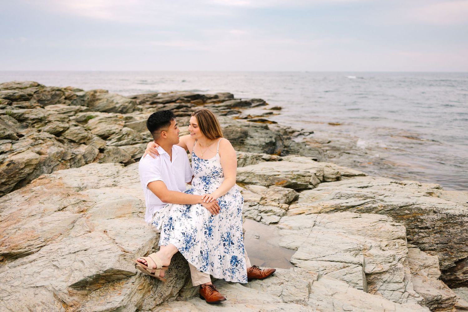 Couple sitting on rocky shoreline for engagement photos, sharing a romantic moment with ocean waves on Ocean Drive in Newport Rhode Island