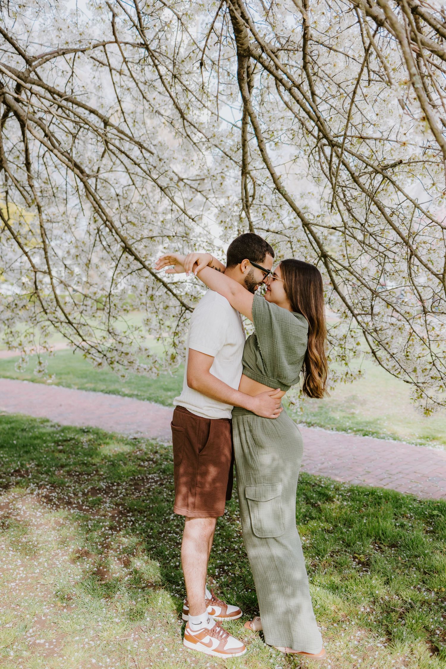 A couple embraces under blooming cherry blossom trees on a spring day along a walking path for engagement photos in Newport Rhode Island