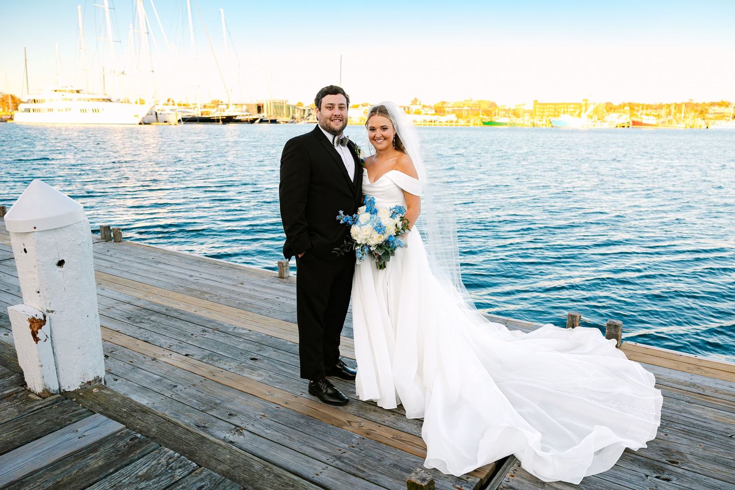 A bride and groom in formal wedding attire poses on a wooden dock with sparkling blue water and boats in the background in Newport RI