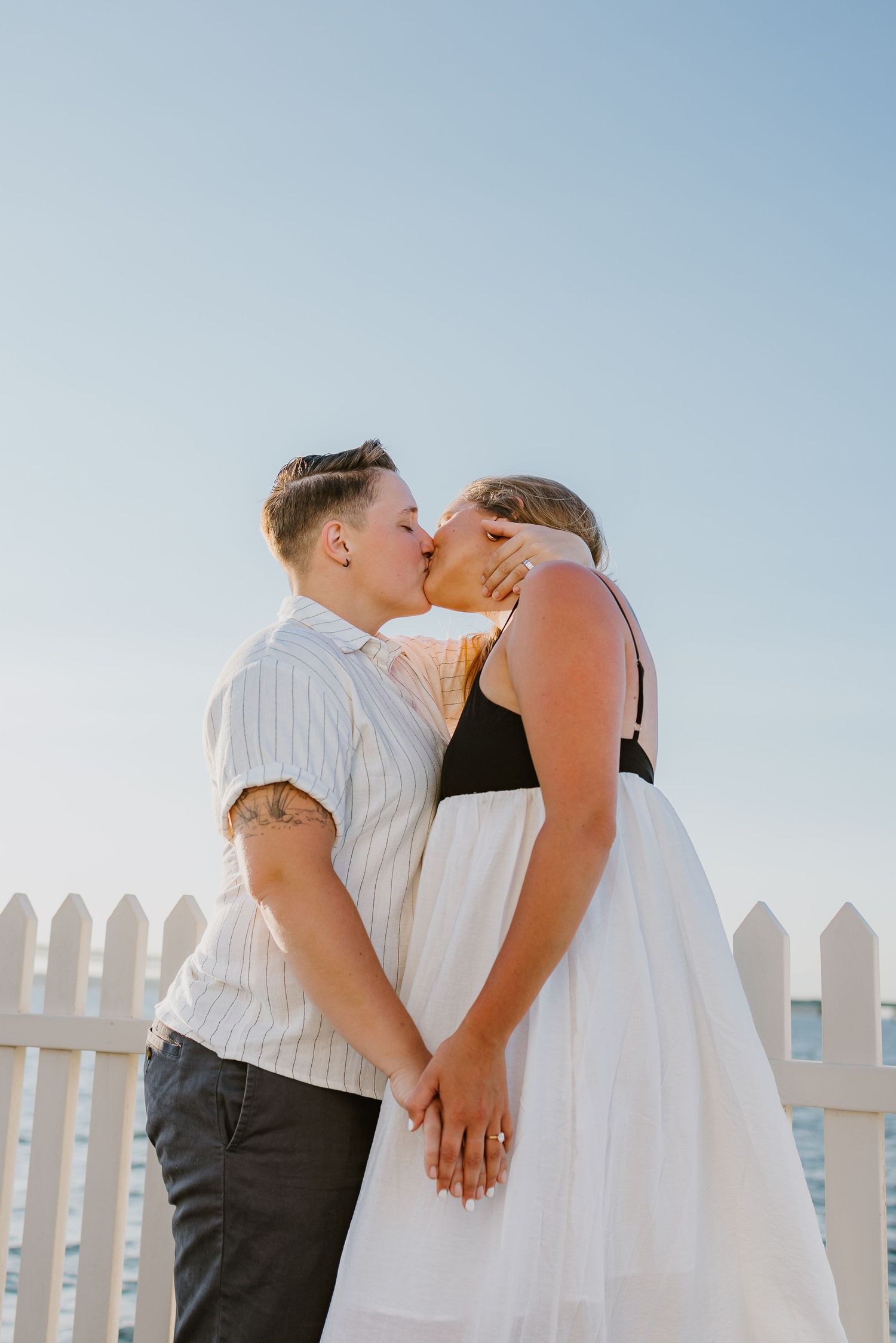 A romantic LGBTQ couple shares a tender kiss at sunset golden hour after surprise proposal at Newport Harbor Island Lighthouse in RI