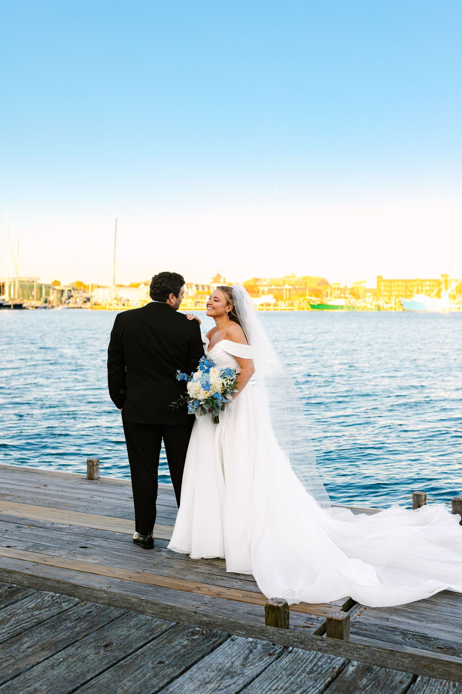 A couple in wedding attire stands on a wooden dock overlooking a waterfront cityscape at sunset.