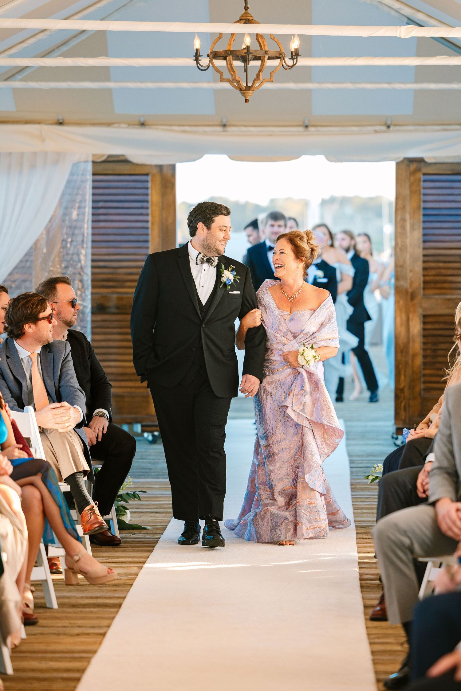 Groom and his mother at an elegant wedding ceremony in a rustic wooden venue with white drapes and chandeliers.