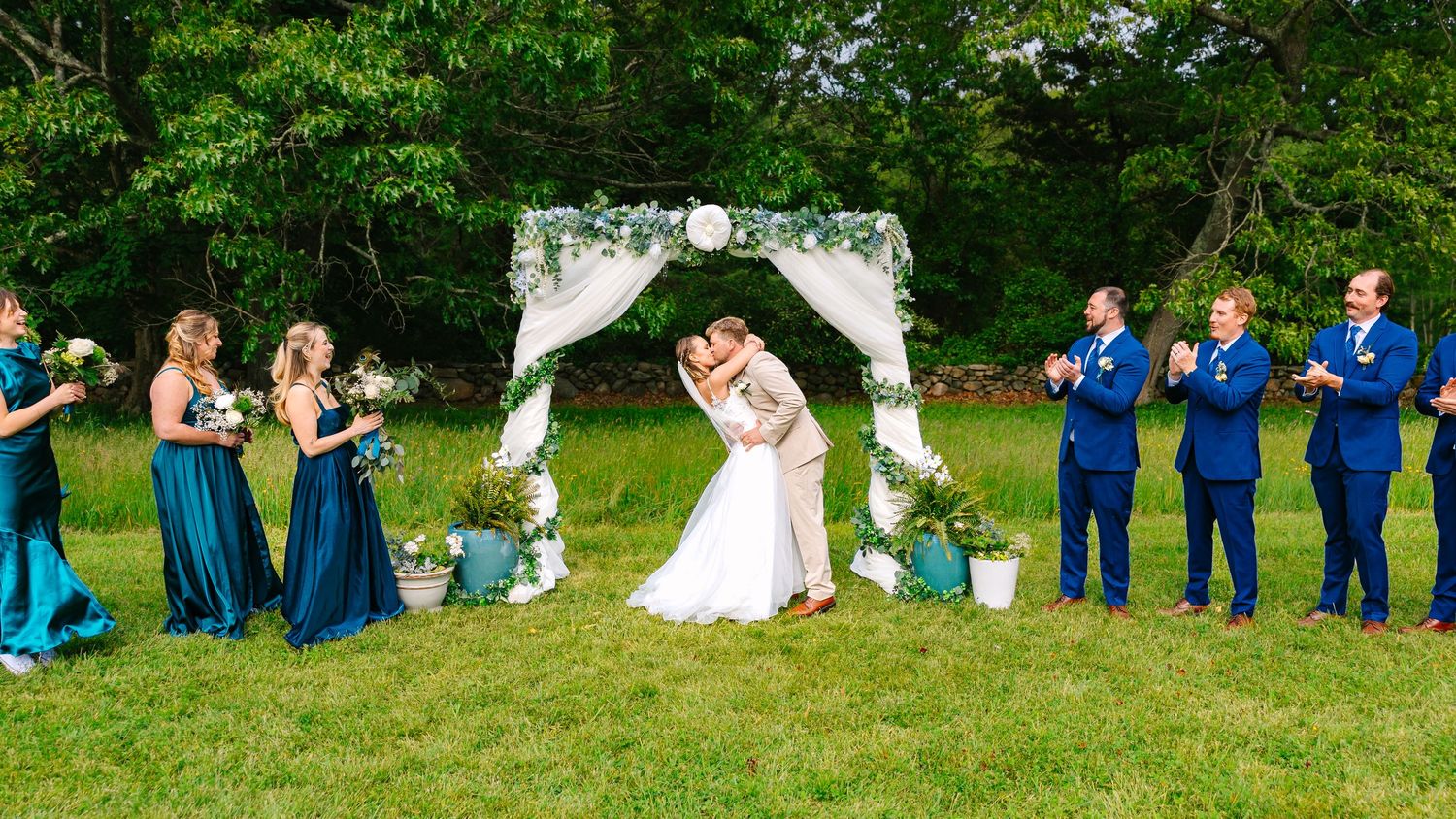 Wedding party in blue attire poses on green grass during outdoor summer celebration.