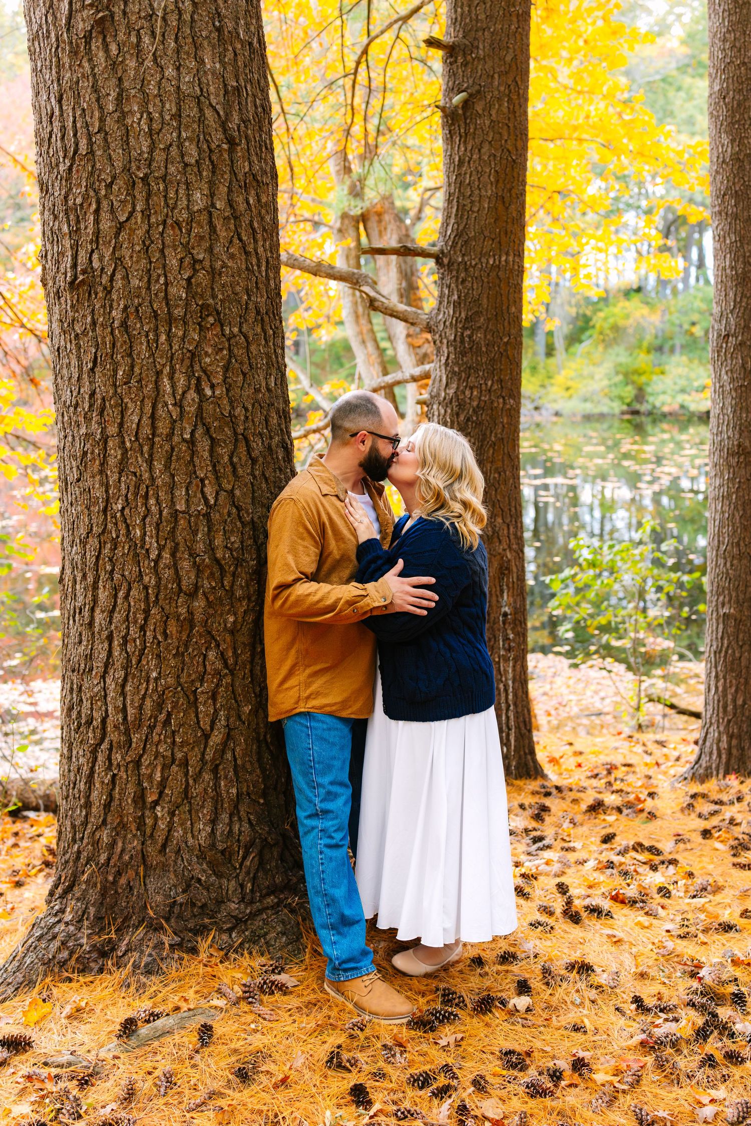 A couple shares an intimate moment against a tree trunk during autumn engagement photos at Goddard State Park in Warwick Rhode Island