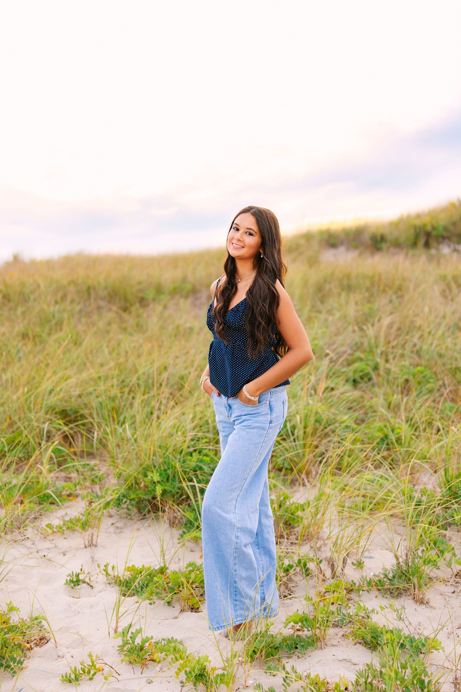 A high school senior in a blue shirt poses on sandy dunes with tall grass during a summer golden hour sunset at Horseneck Beach in MA