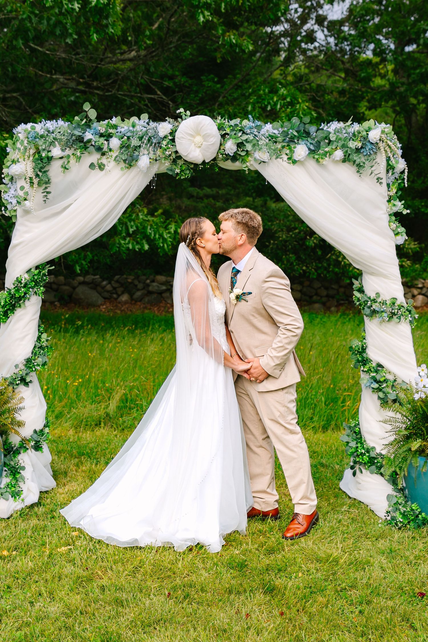 Romantic outdoor wedding photos on green lawn with flowing white fabric drapes and greenery decorations creating a natural archway.