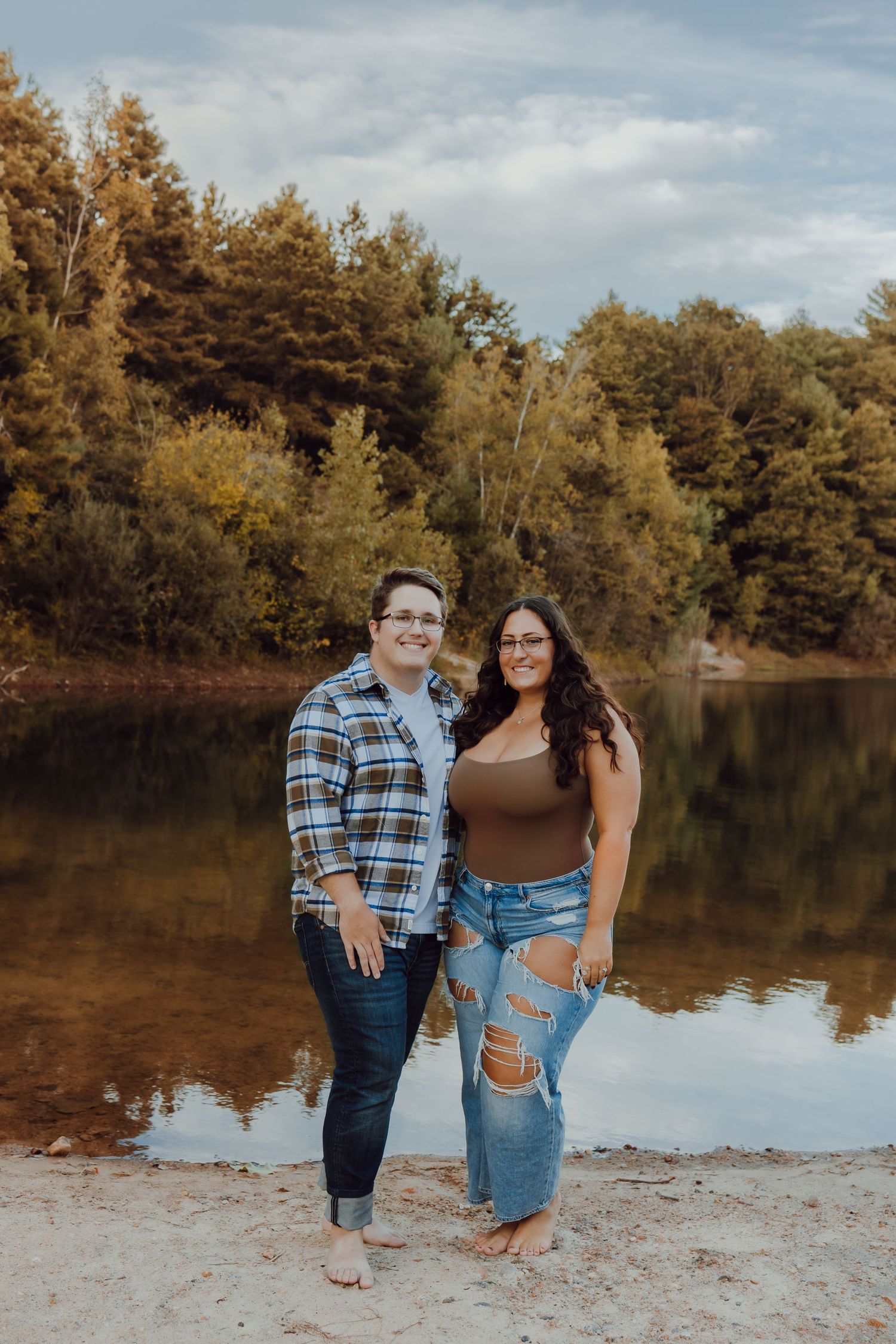 A couple poses together by a serene lake with autumn trees reflecting in the calm water during golden hour in West Greenwich Rhode Island