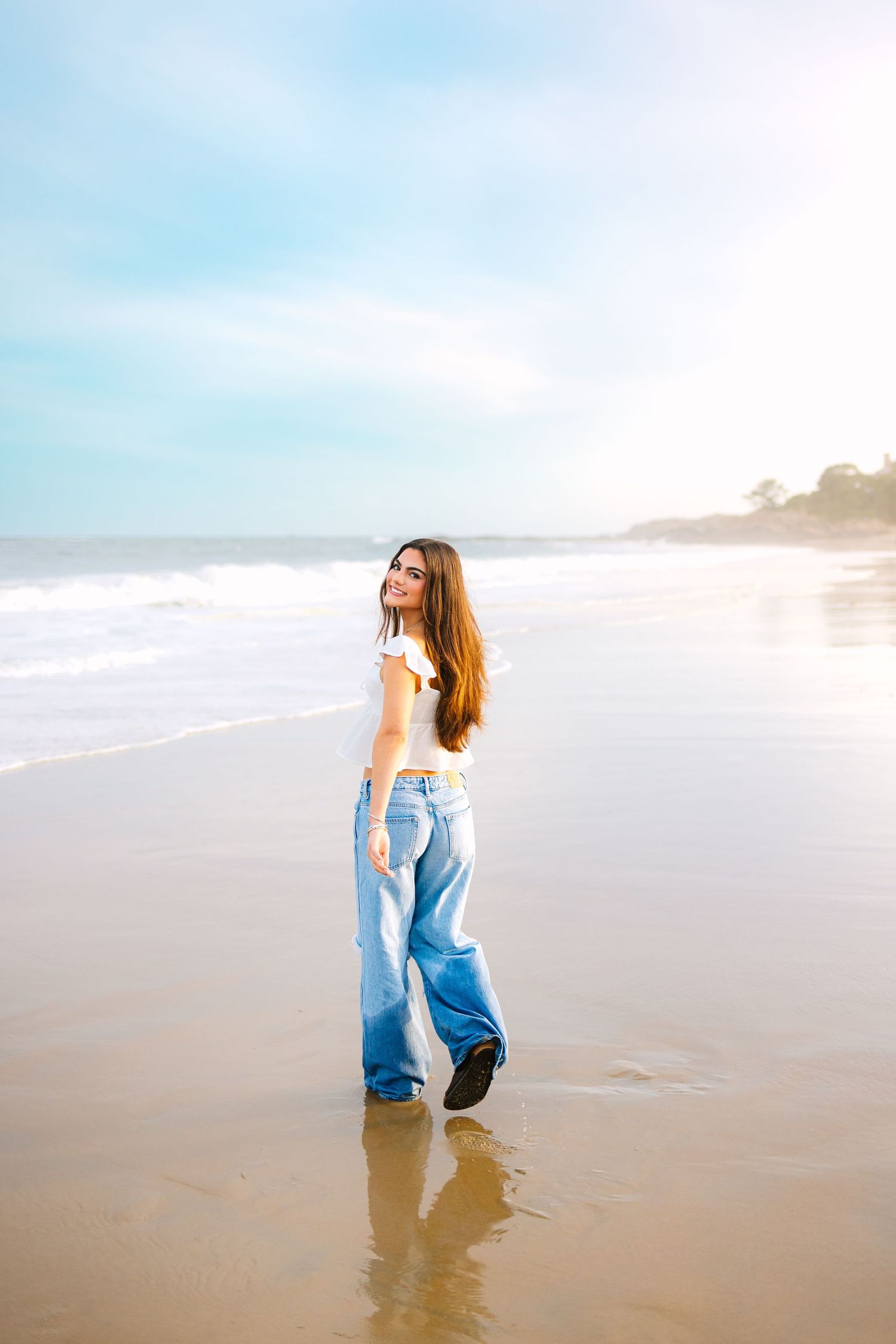 A high school senior walks candidly by the misty shore for senior photos at sunset golden hour at a beach in Boston Massachusetts
