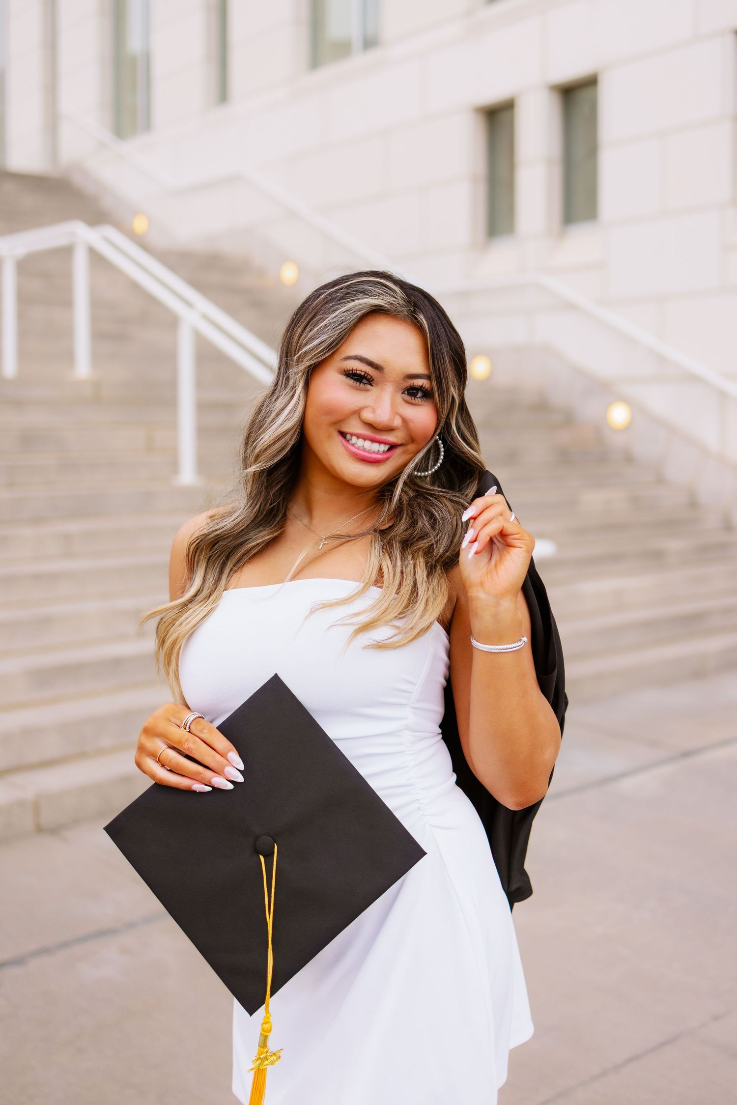 A graduate in a white dress and black graduation gown holds their cap while posing on concrete steps at UMASS Boston Massachusetts
