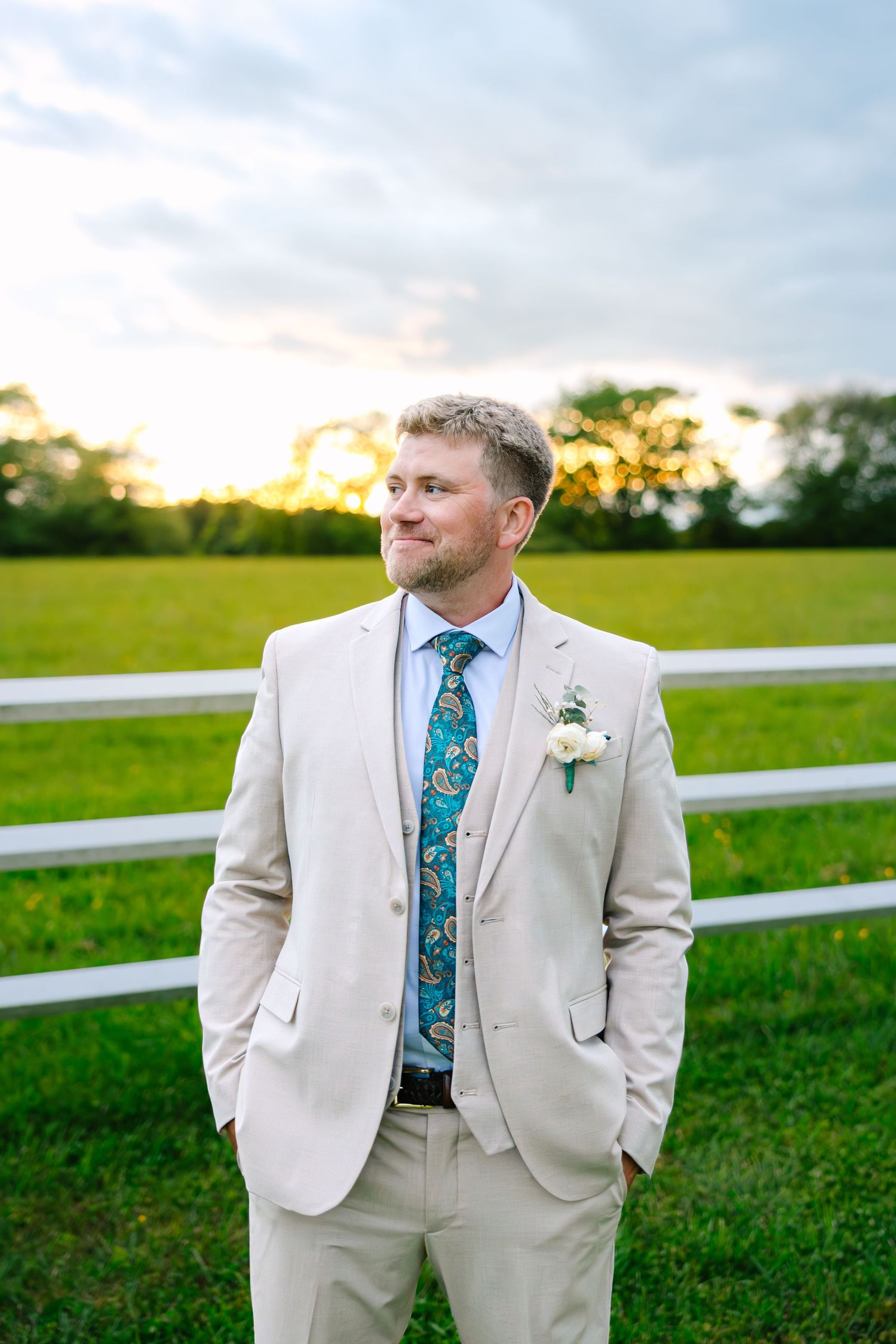 A groom in a tan suit with a floral tie stands in front of a white fence at sunset in a grassy field.
