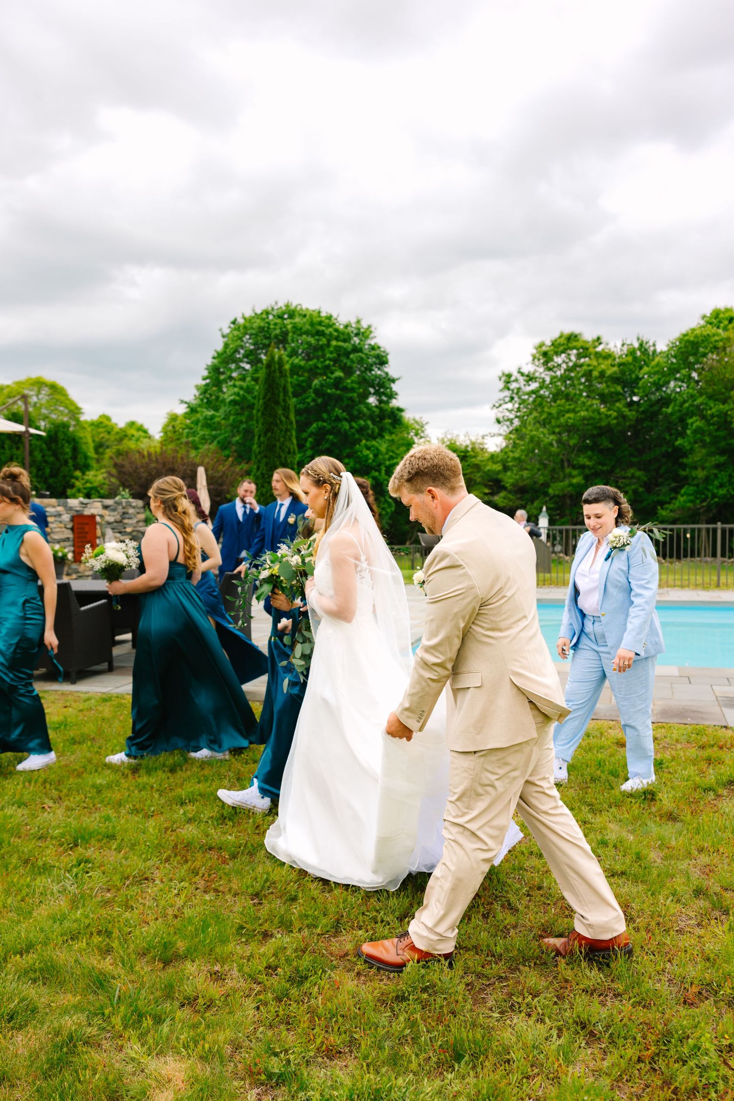 A playful bridal party in blue and beige attire walks candidly outdoors on a grassy lawn.