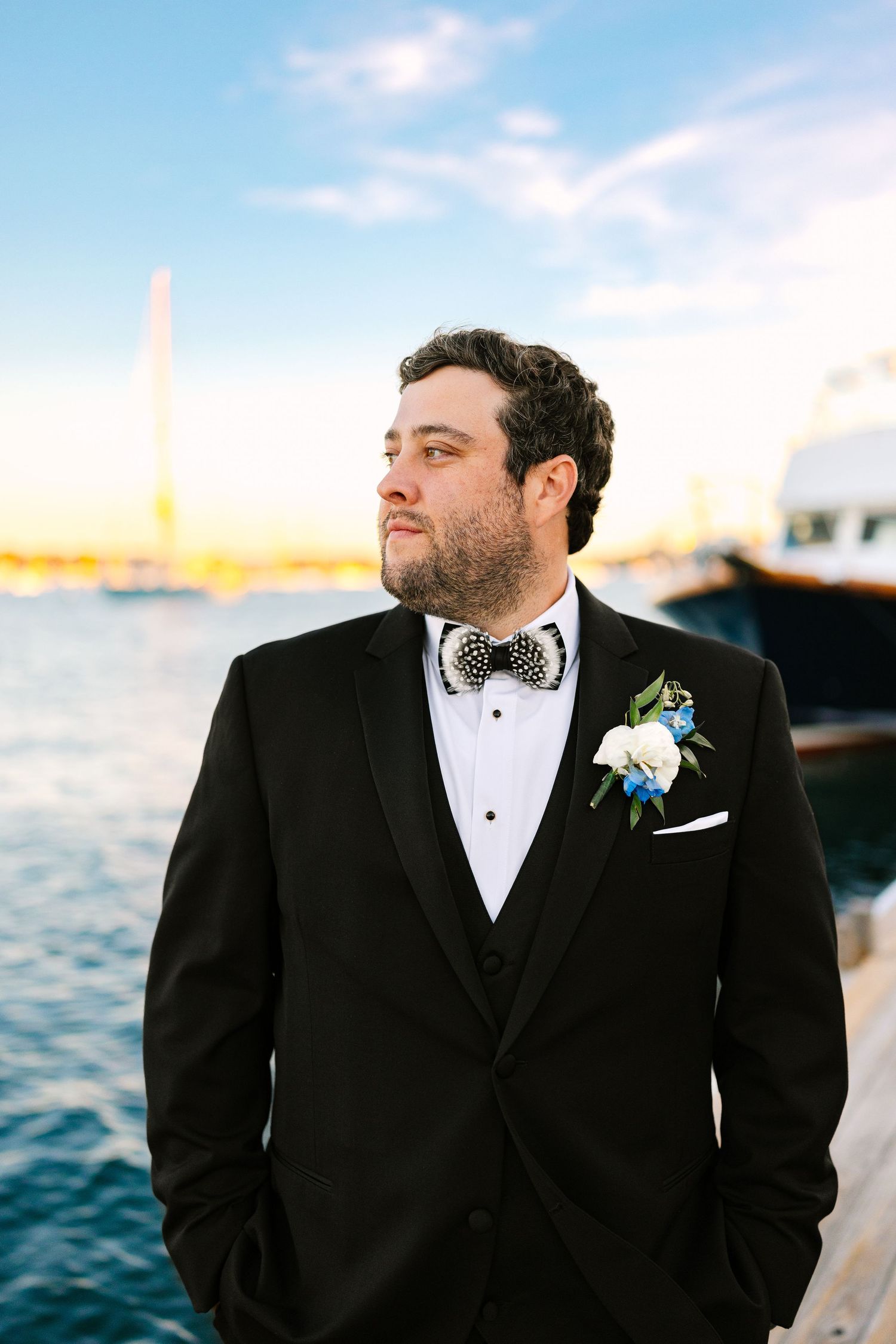 A groom in a black tuxedo and bowtie poses against a waterfront backdrop during sunset at The Regatta Place in Newport, Rhode Island