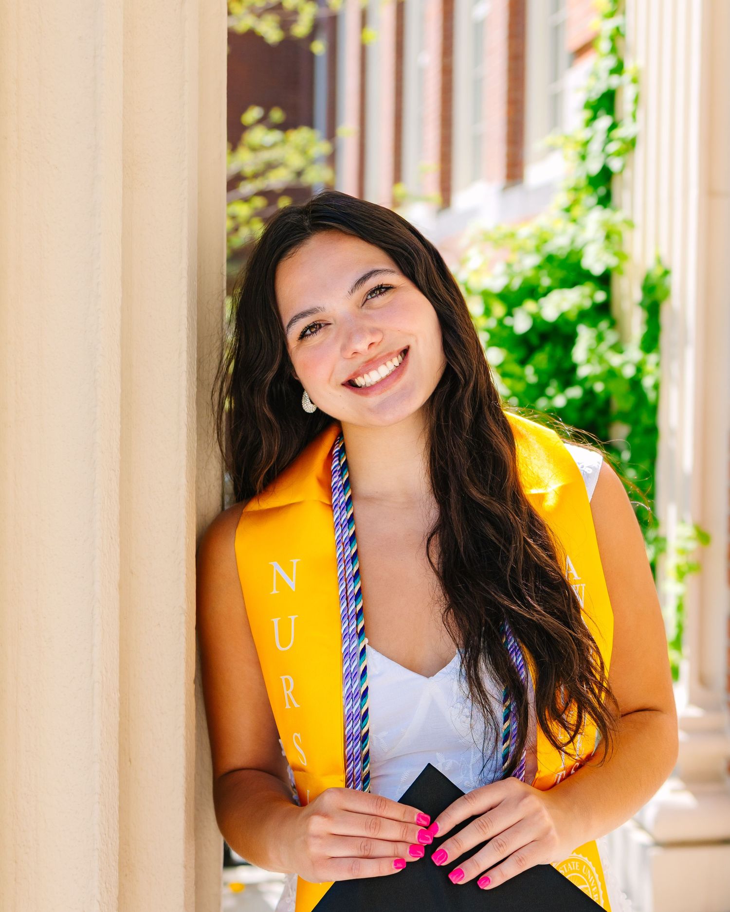 A graduate wearing a yellow stole posing for graduation photos by school building at Worcester State University in Worcester, Massachusetts