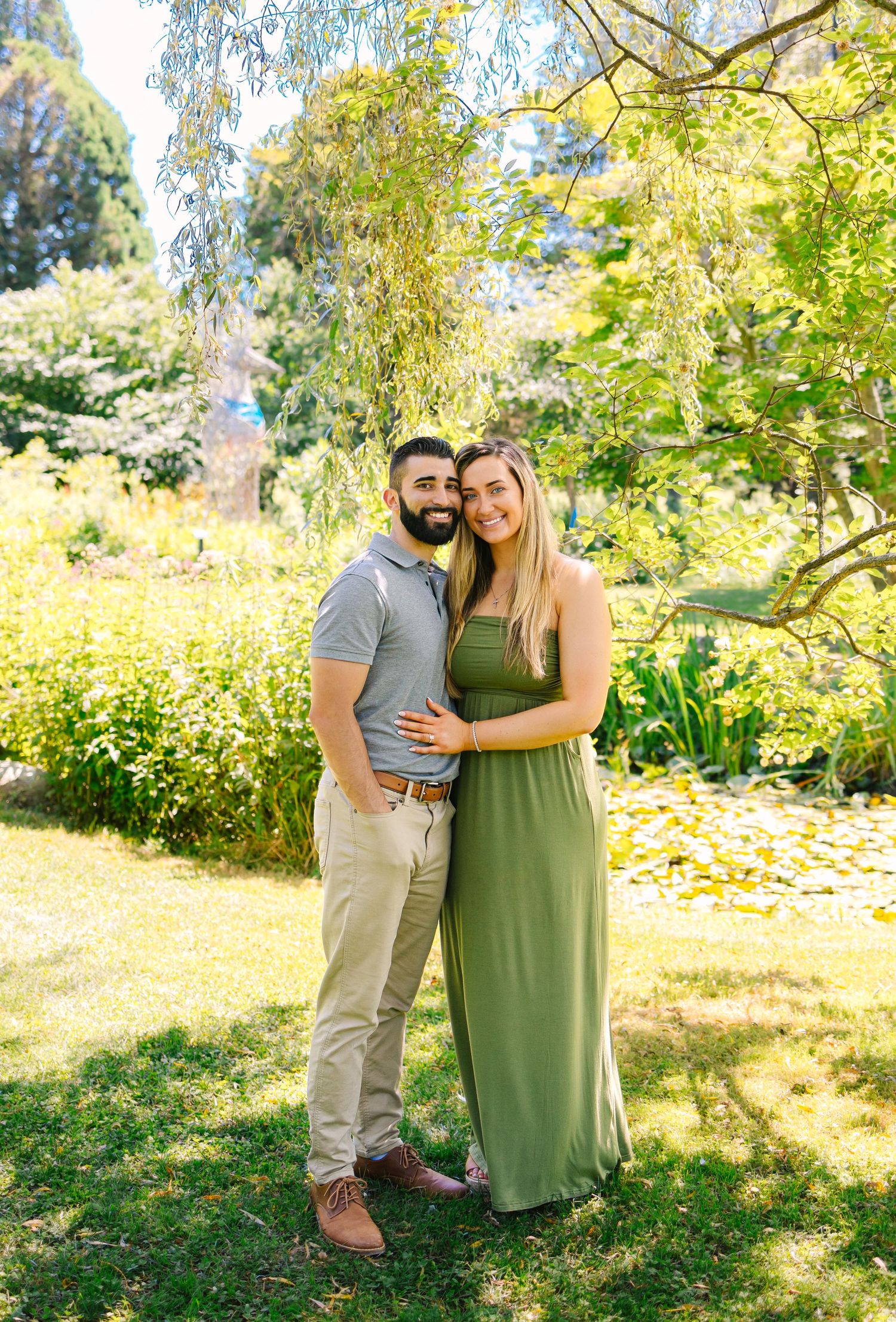 A couple embraces while standing in a sunny garden with lush greenery and yellow flowers in the background.