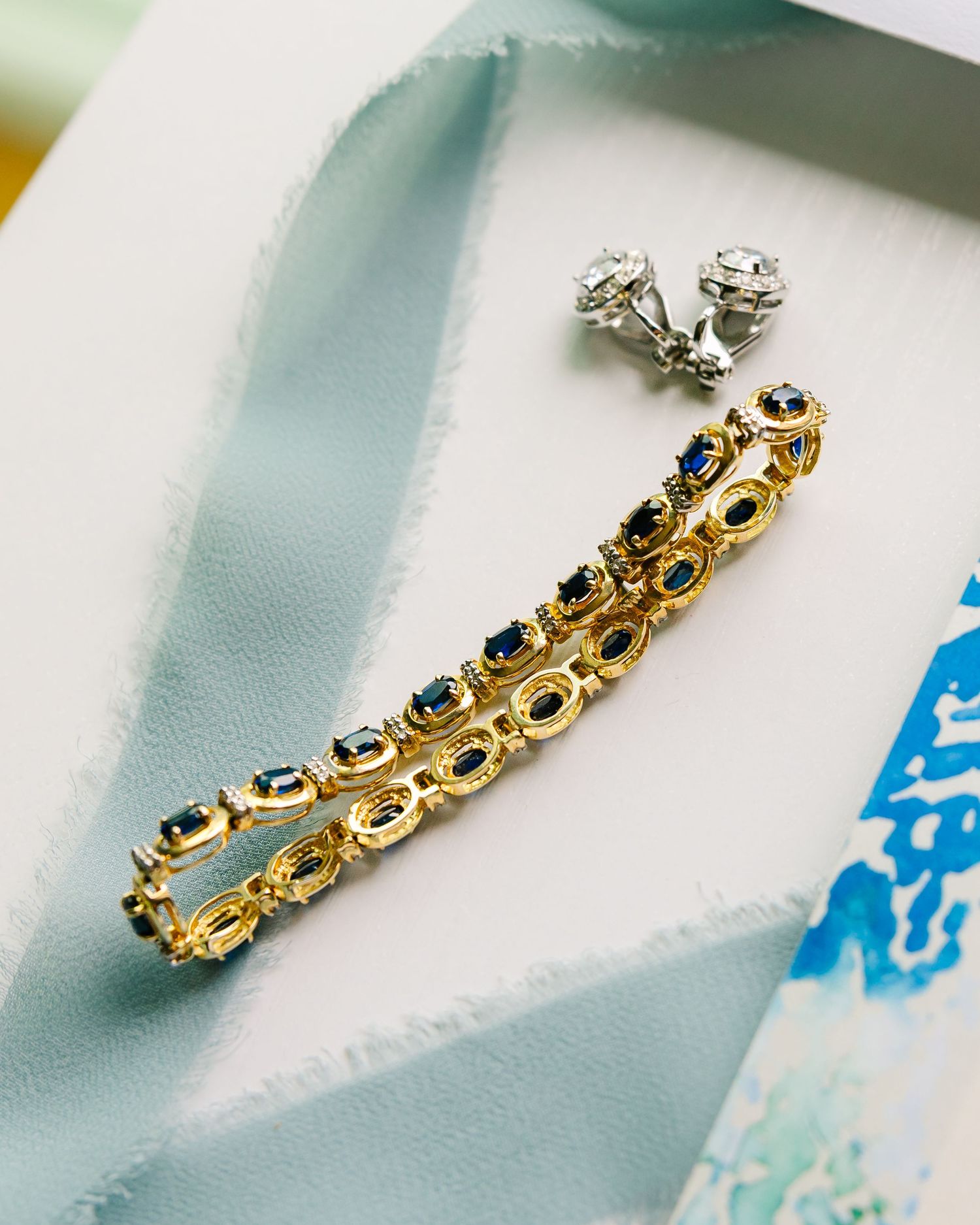 Elegant gold bracelet with blue gemstones and matching diamond stud earrings displayed on light blue fabric.