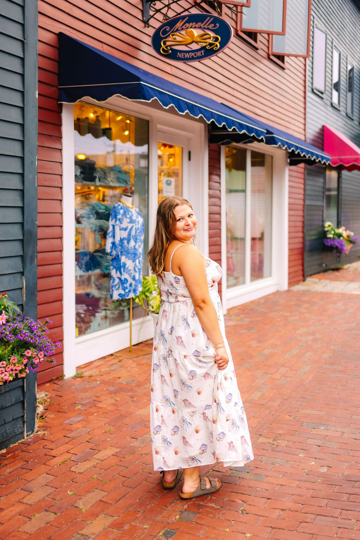 A high school senior in a floral dress walks along a brick sidewalk past storefronts in a shopping district at Bowen's Wharf in Newport RI