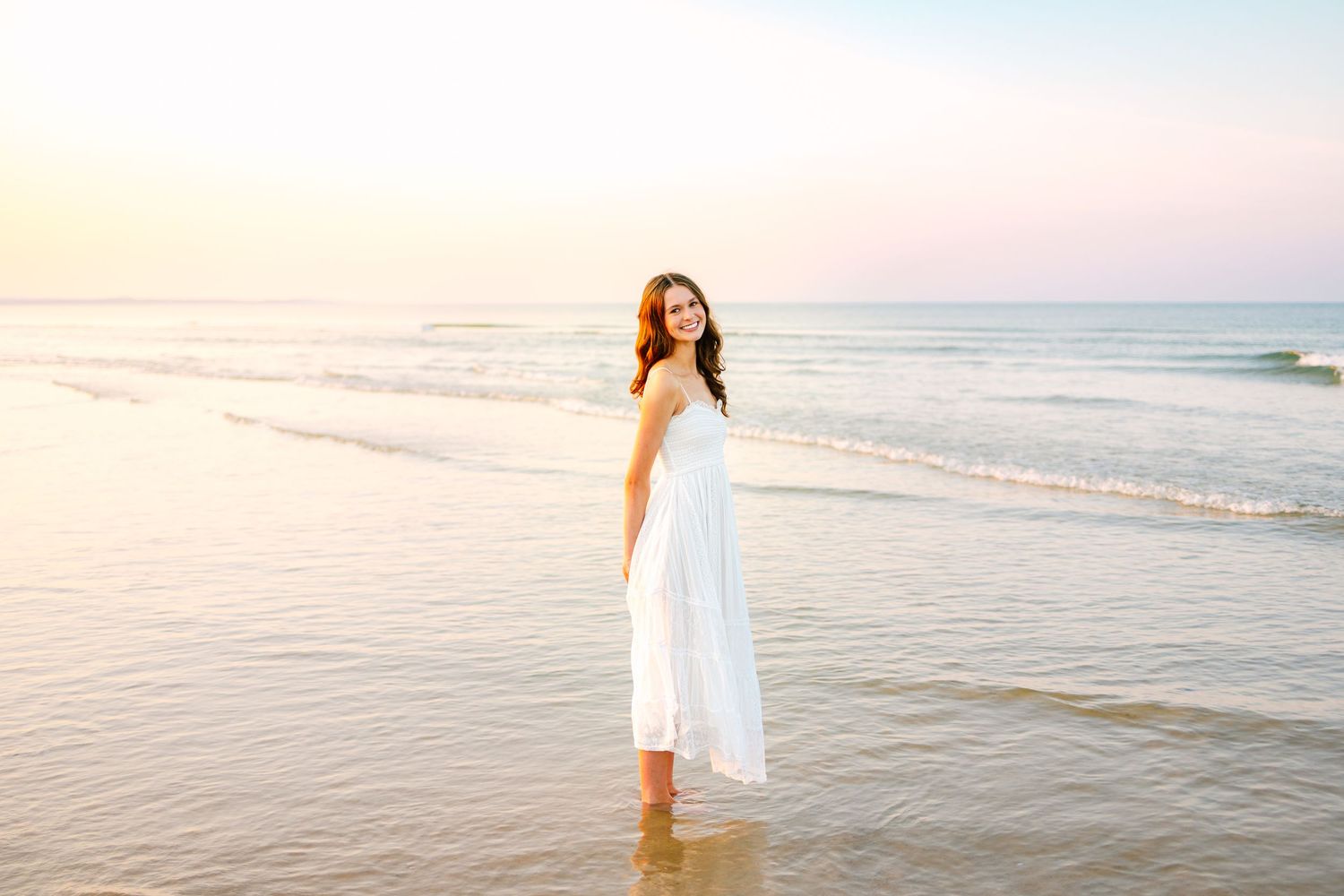 A high school senior stands in shallow ocean water during a dreamy sunset golden hour senior photos at Scusset Beach in Sagamore MA