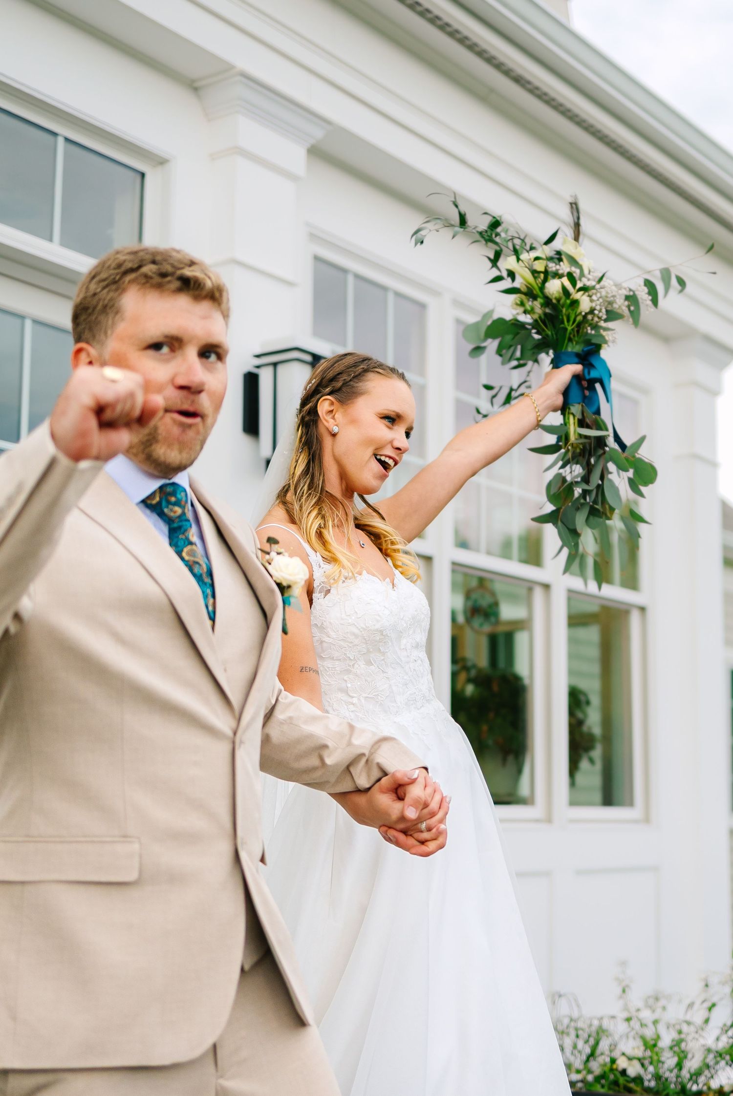 A couple in wedding attire dances joyfully together outside a white building with large windows.