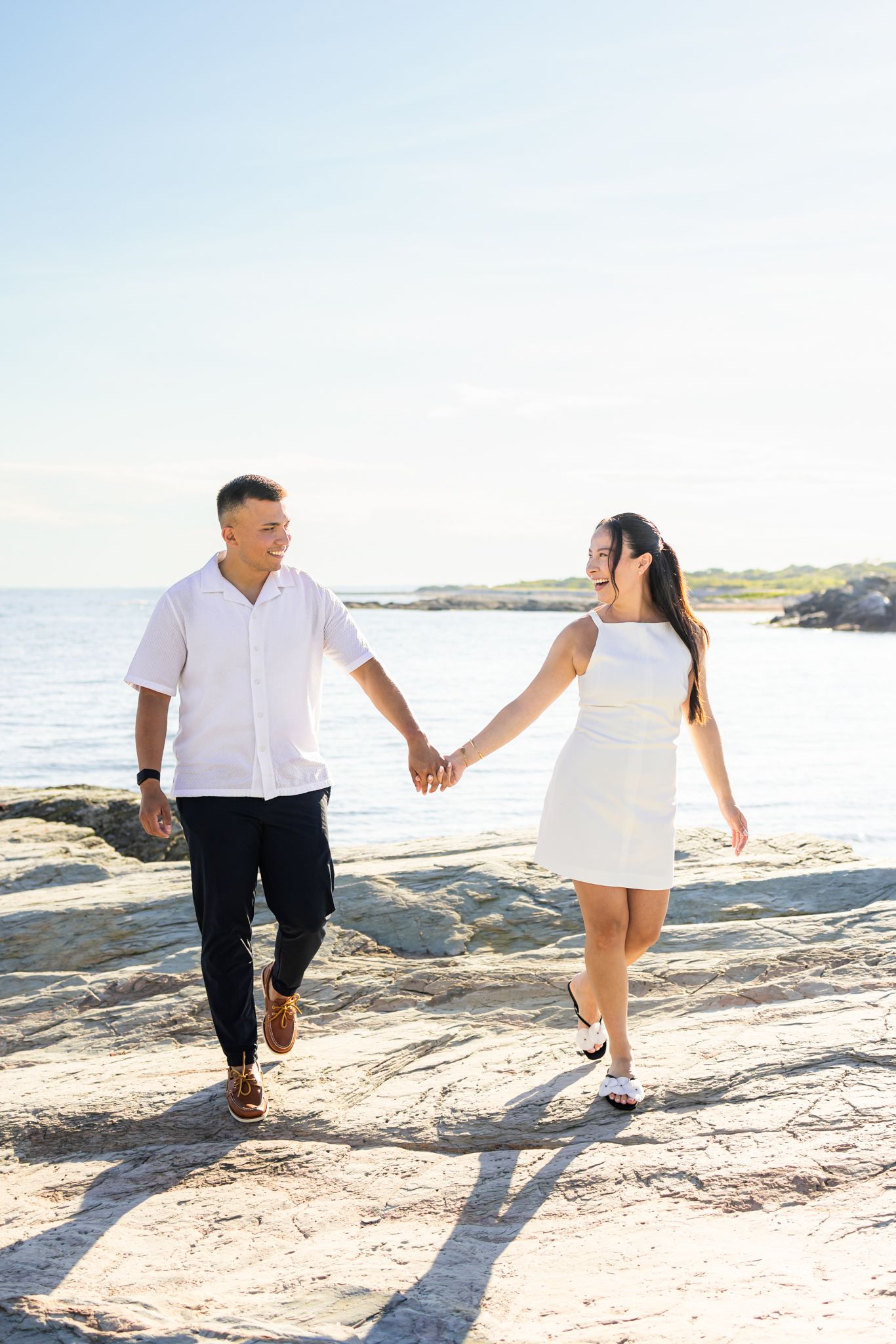 A couple holds hands while walking along a rocky cliff coastline after surprise proposal at sunset golden hour at Ocean Drive in Newport RI