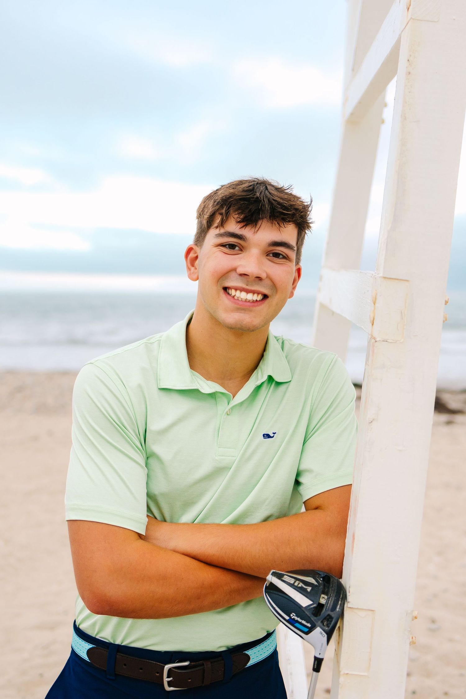 High school senior in a mint green polo shirt smiles while posing by a lifeguard chair on a cloudy day at Horseneck Beach in Westport MA