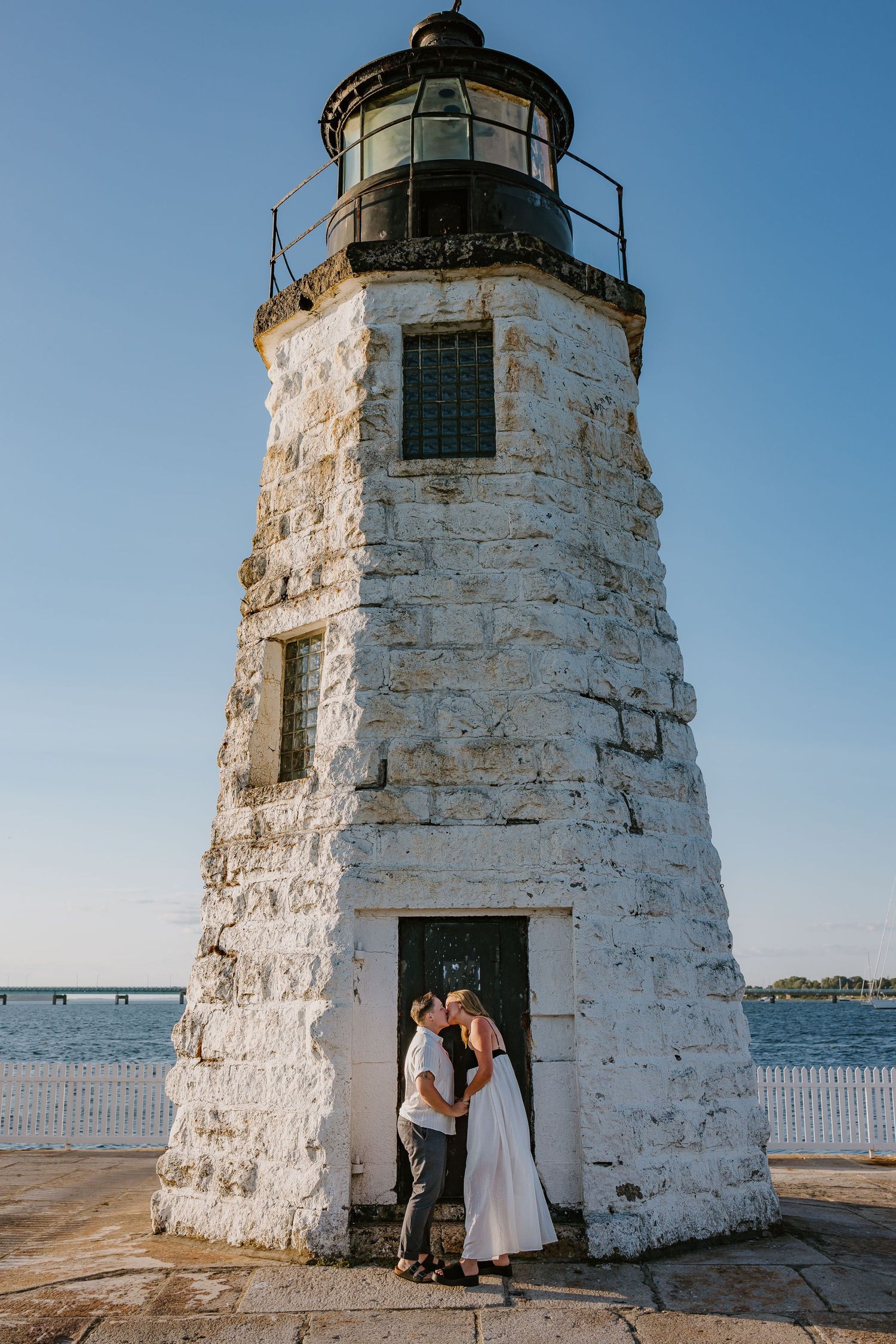 A LGBTQ couple kisses in front of Newport Harbor Island Lighthouse during a summer sunset in Newport Rhode Island