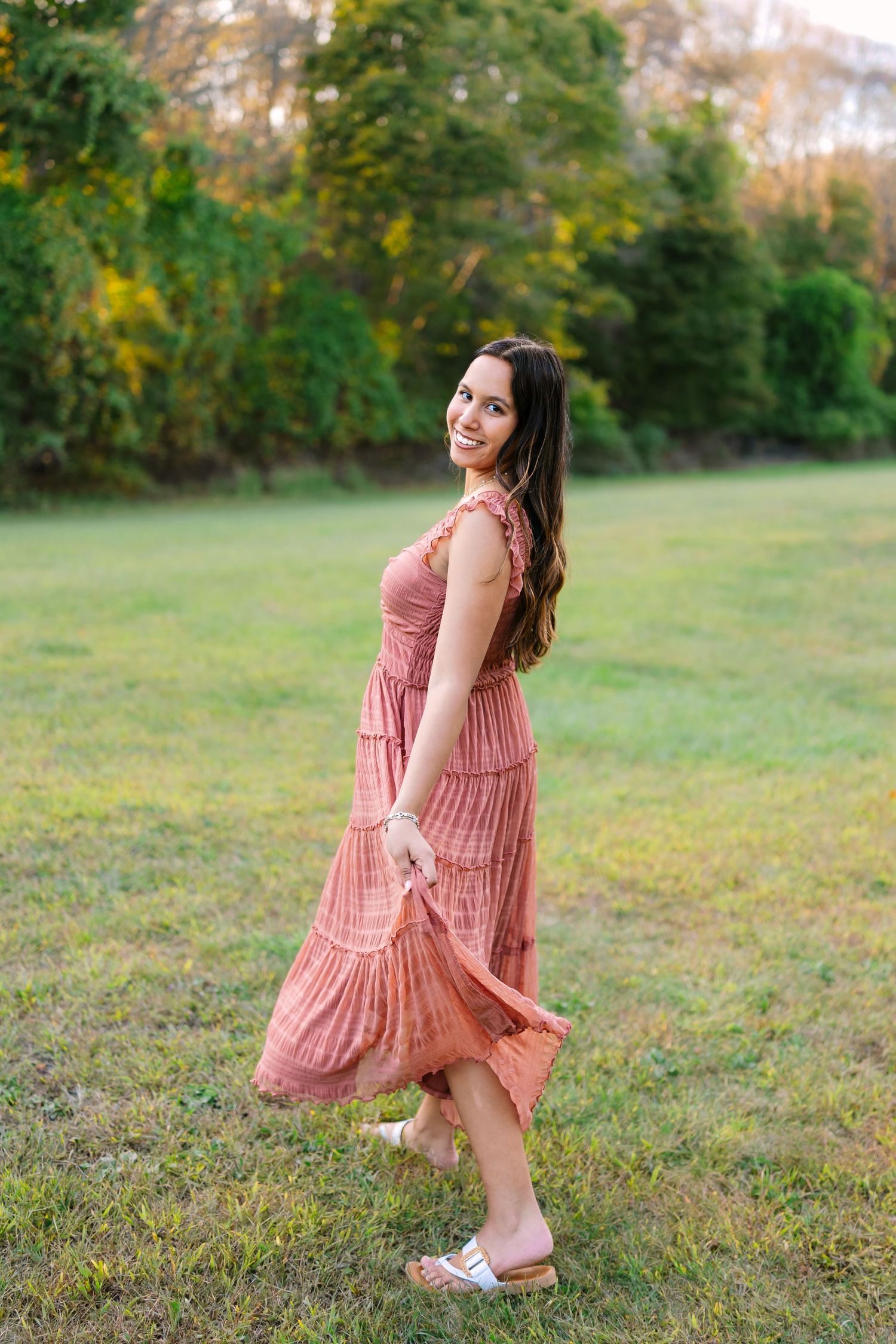 A high school senior in a flowing pink dress twirls playfully in a grassy field during golden hour in the autumn at Colt State Park in RI