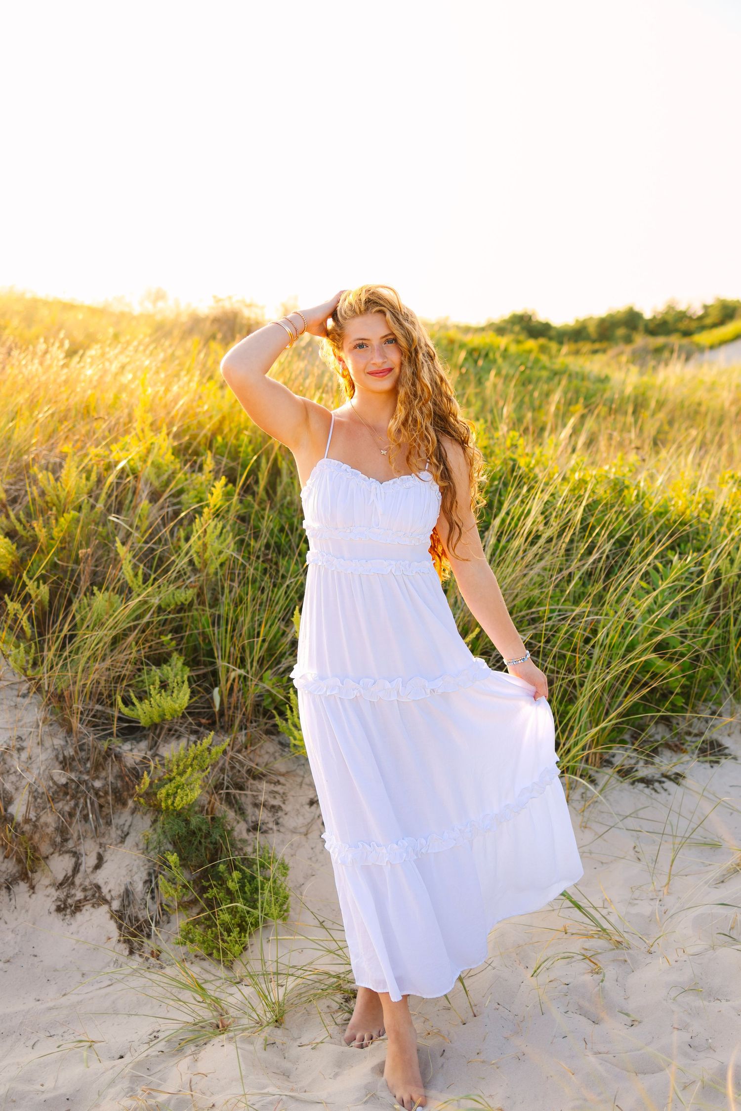 A high school senior in a flowing white sundress dances gracefully on a sandy beach at sunset among tall sea grass at Horseneck Beach in MA