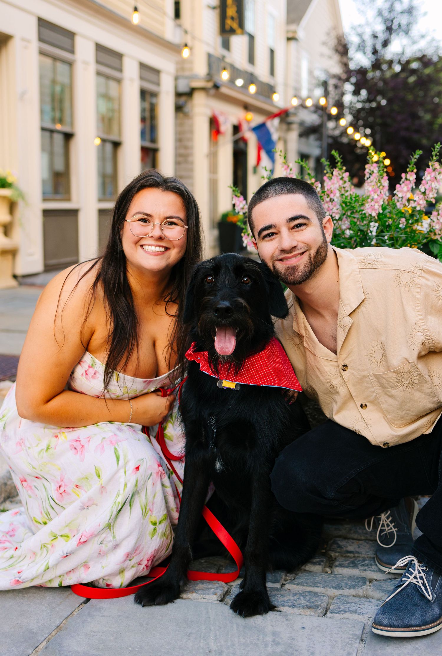 A happy black Labrador wearing a red bandana sits outdoors with couple on stone steps near flowering plants in Downtown New Bedford MA