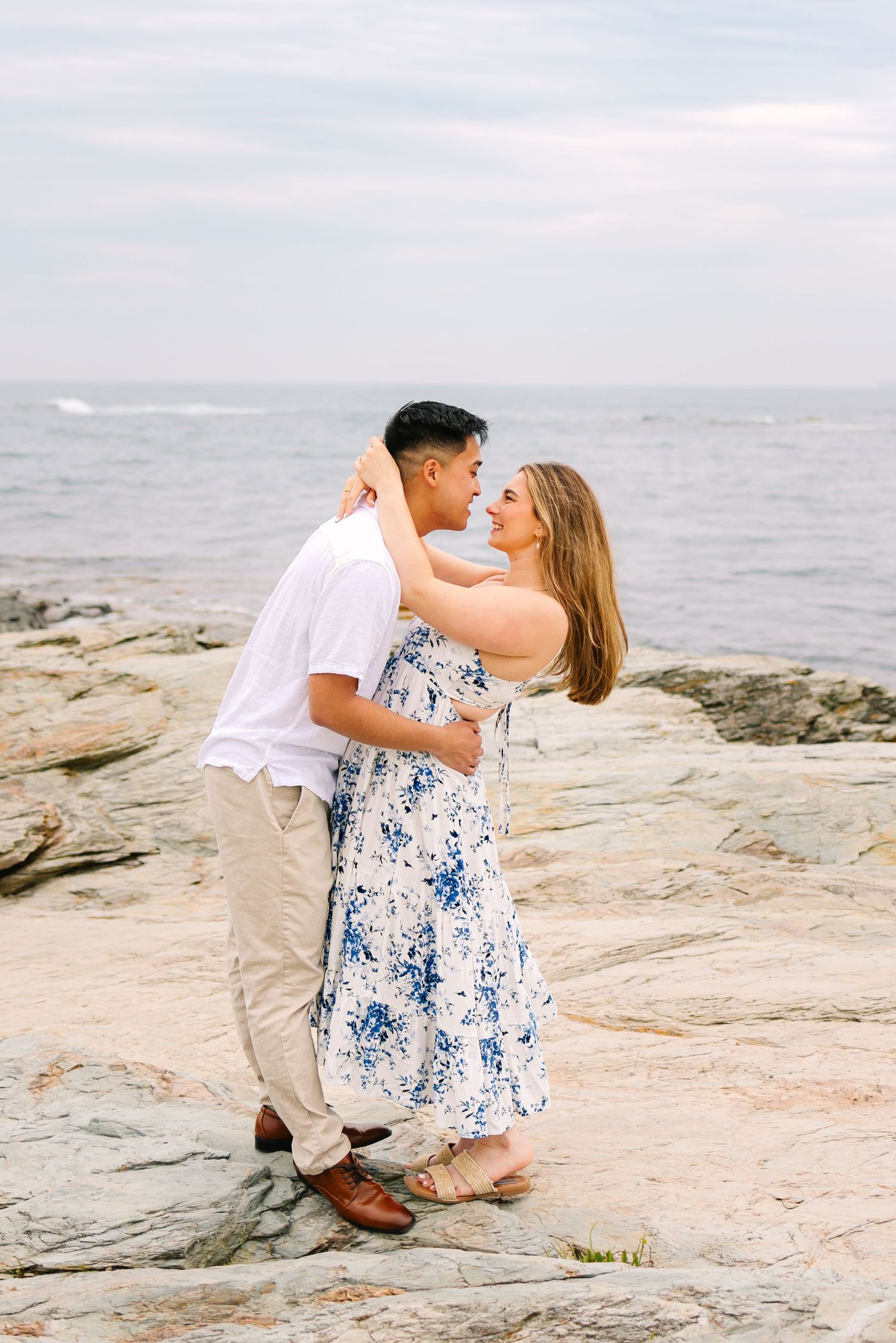 A romantic couple embraces on rocky coastal cliffs overlooking the ocean on a misty afternoon for engagement photos in Newport Rhode Island.