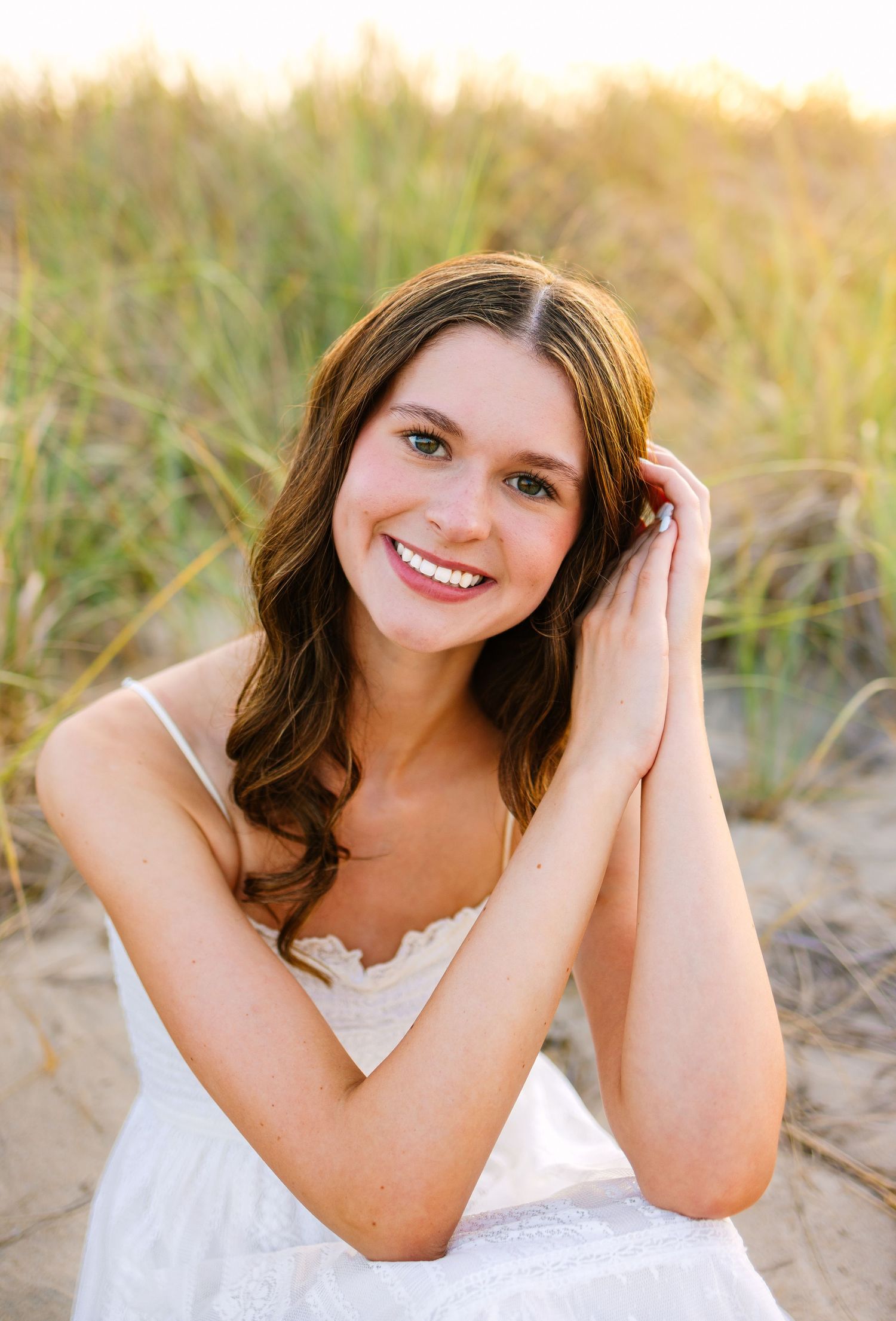 A portrait of a high school senior in a white dress smiling at sunset with tall grass in the background at Scusset Beach Cape Cod MA