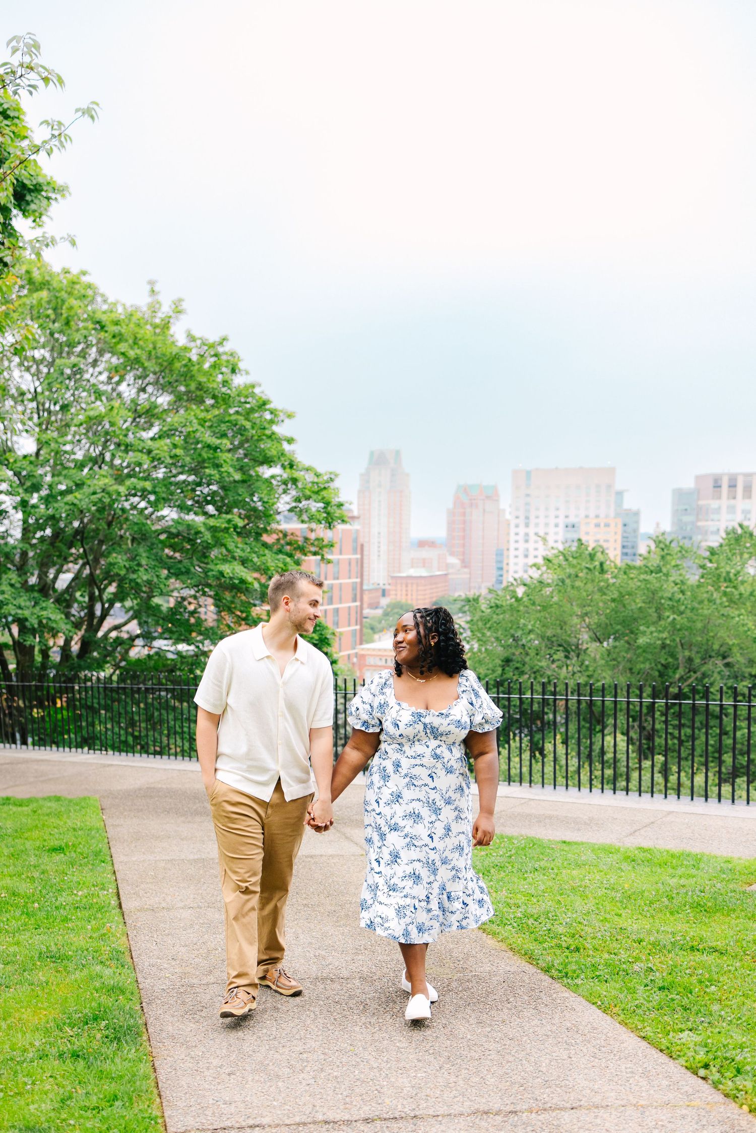 A couple takes a romantic stroll along a paved path in a park with the Providence city skyline visible in the background.
