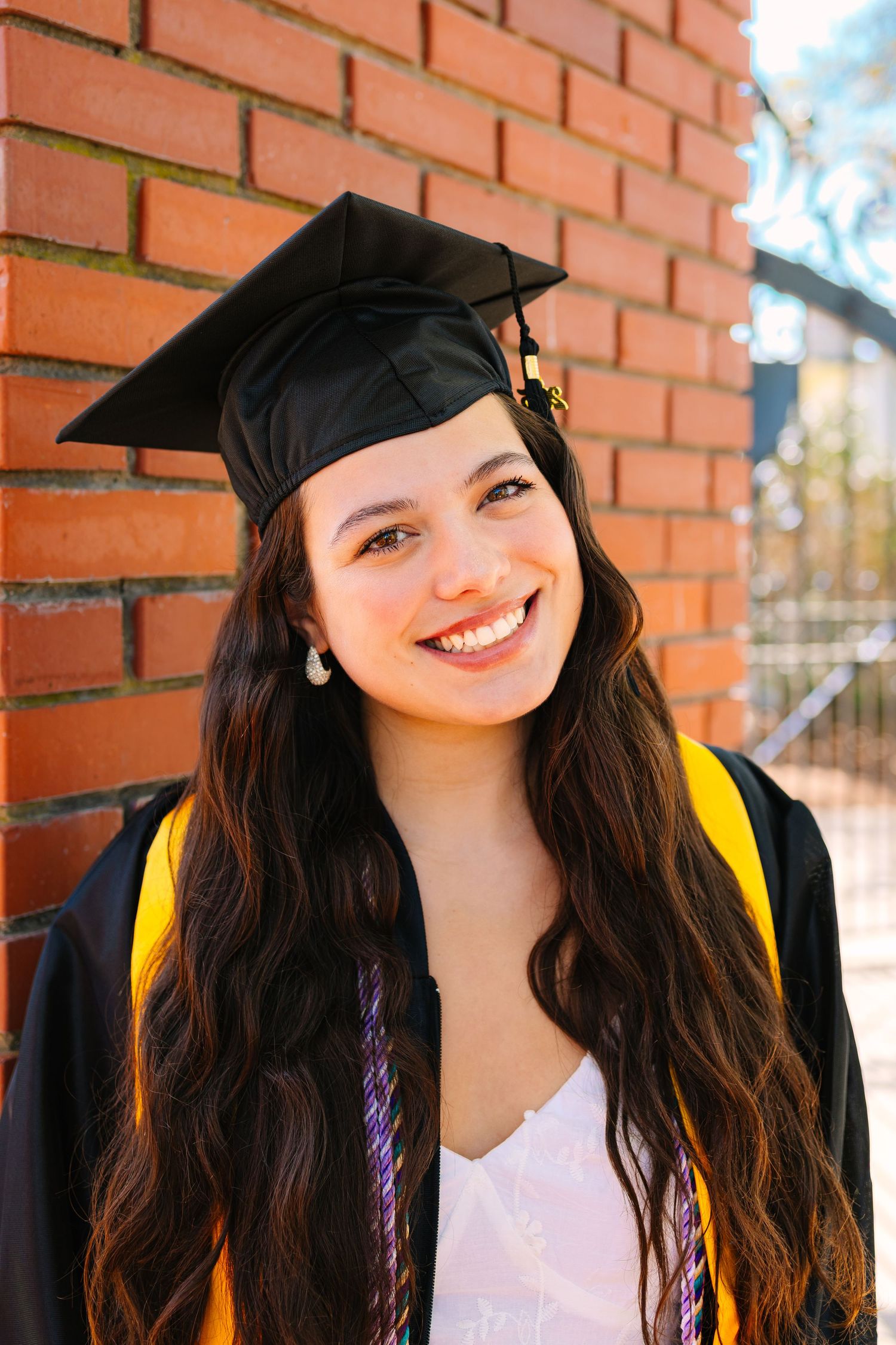 Graduate in cap and gown smiles brightly against brick wall for graduation photos at Worcester State University in Worcester Massachusetts