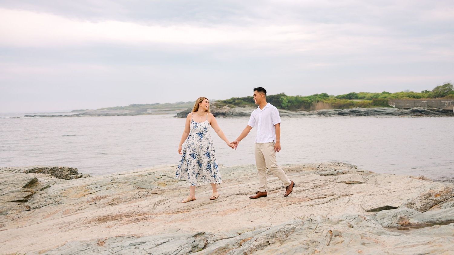 A couple holds hands while walking along rocky coastal shoreline at sunset near the ocean on Ocean Drive in Newport Rhode Island engagement
