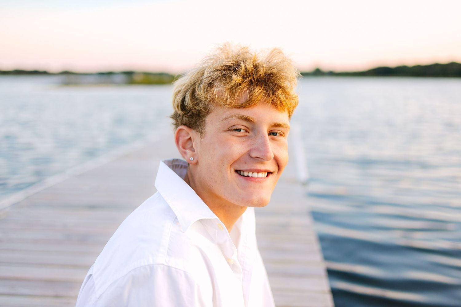 A high school senior in a white button-down shirt smiles while sitting on a wooden dock by a serene lake at golden hour in South Coast MA