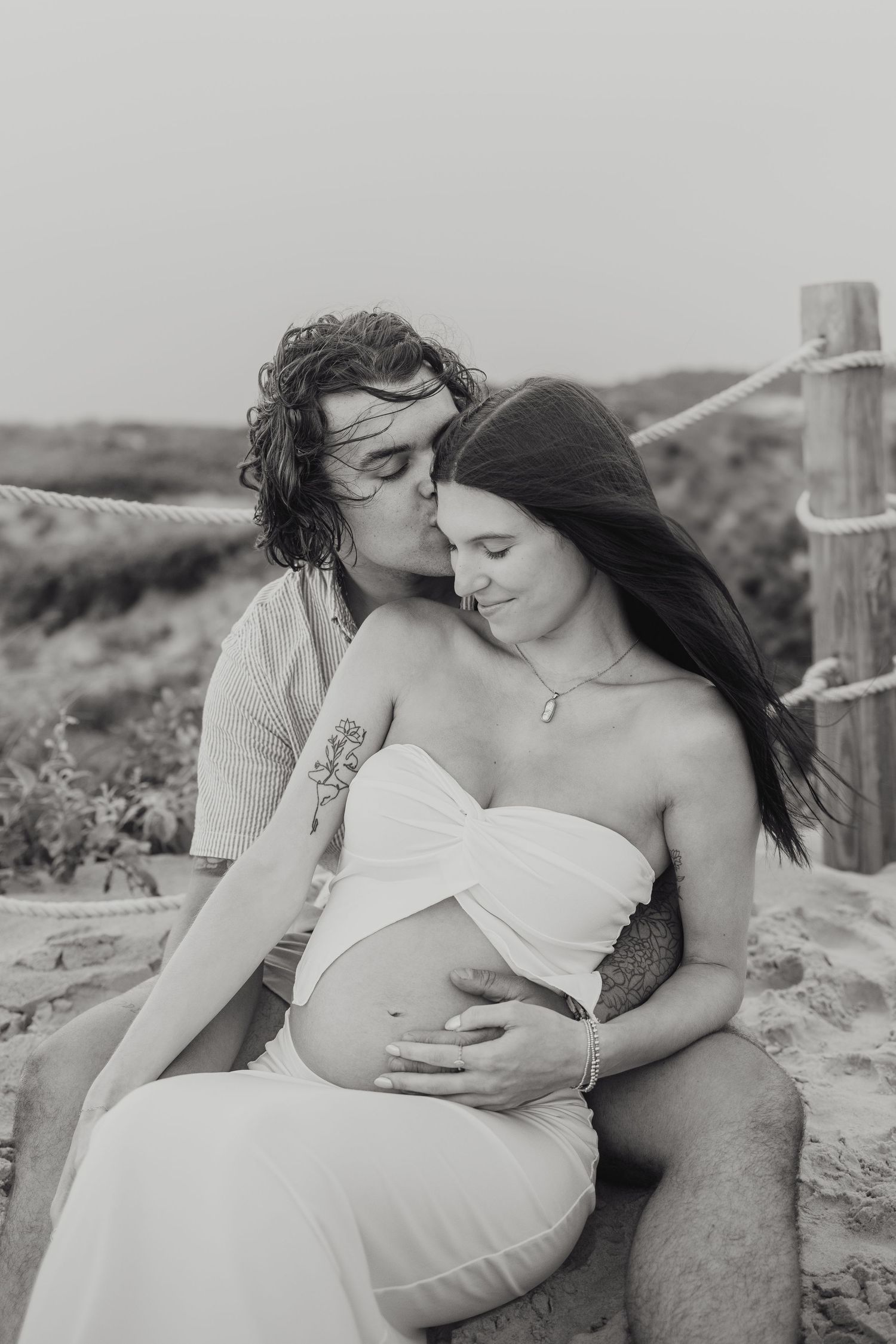 A romantic black and white photo of man embracing his pregnant wife by the sand dunes at Horseneck Beach in Westport, Massachusetts