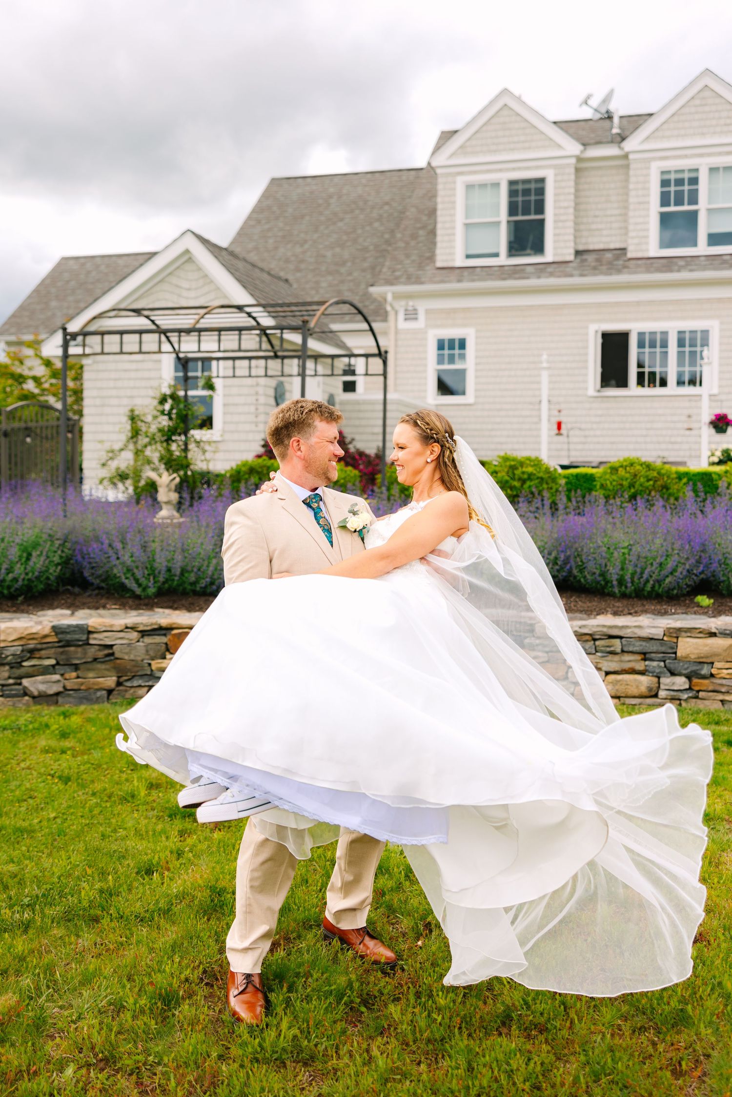 A beautiful wedding couple dances joyfully in front of a white house with purple wildflowers in bloom.