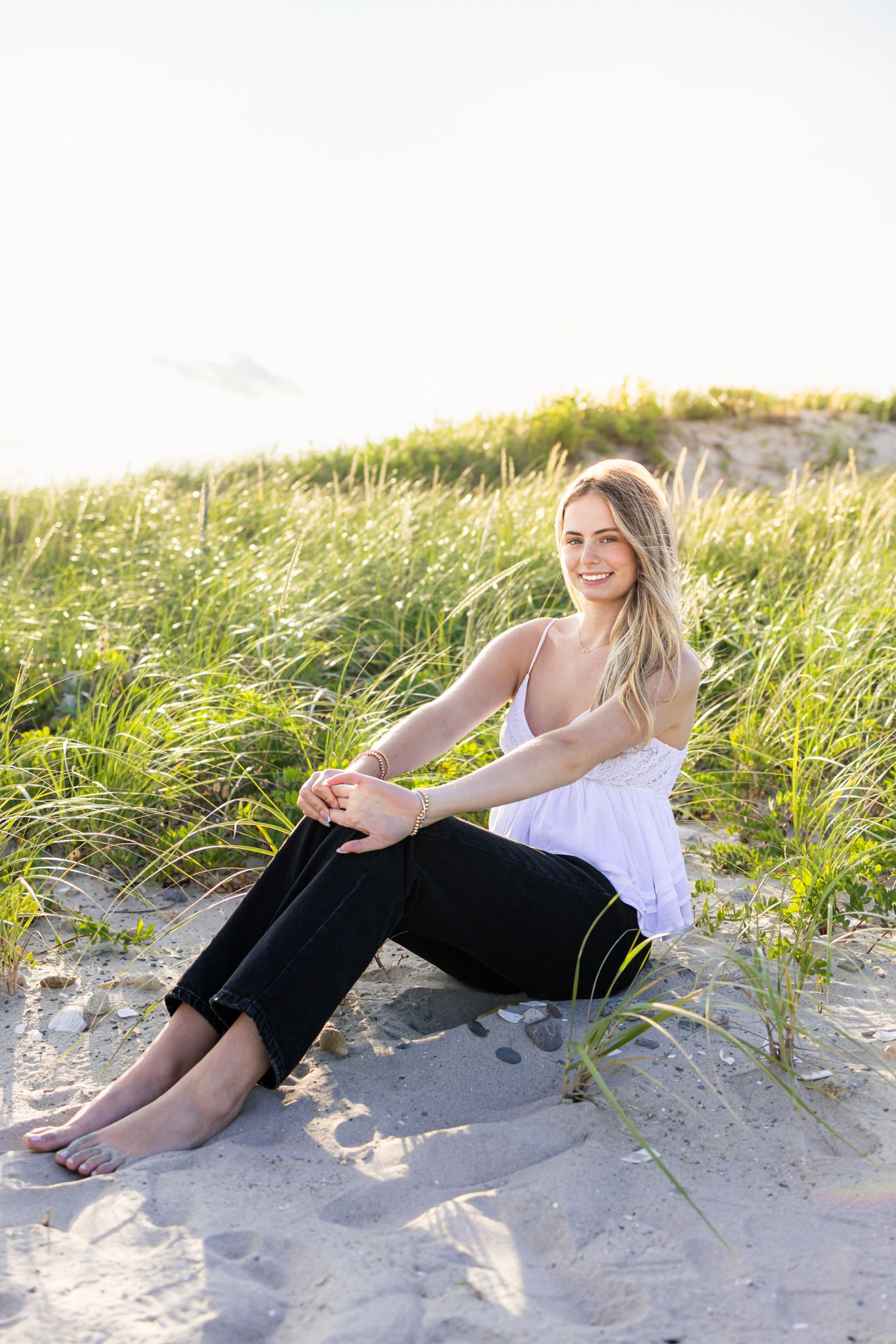 A high school senior sits by the seagrass at sunset golden hour for senior photos at Horseneck Beach in Westport, Massachusetts
