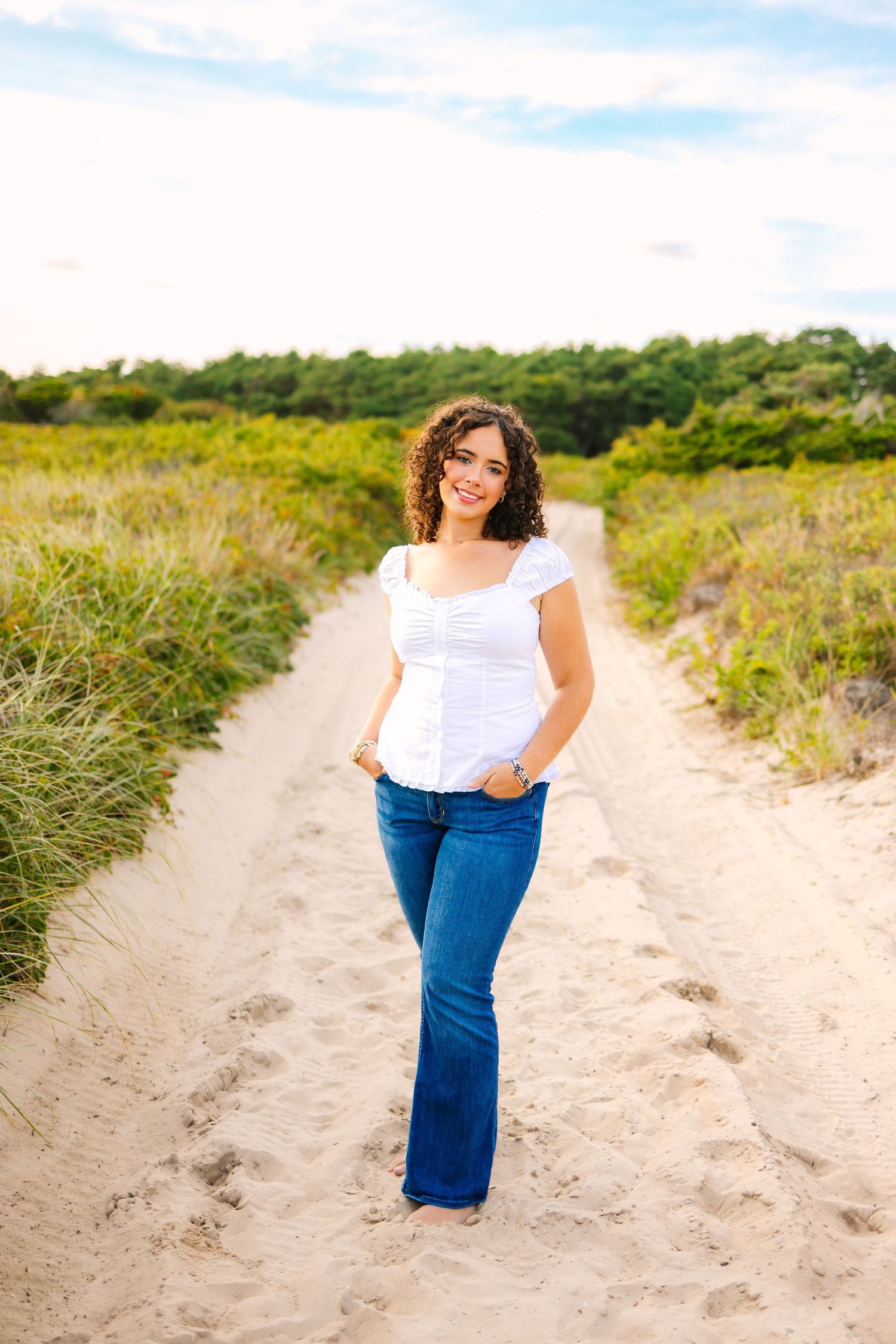A high school senior poses along a sandy beach path surrounded by coastal vegetation at sunset golden hour at Horseneck Beach Westport MA