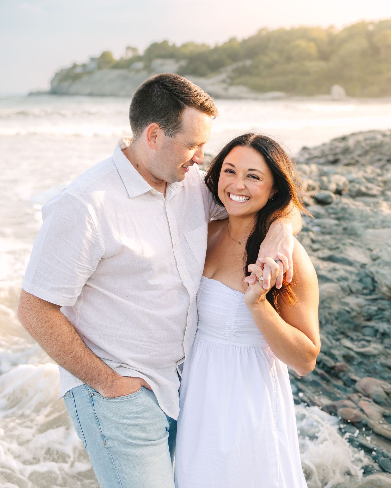 A couple shares intimate moments on a rocky beach at sunset in soft, dreamy lighting.