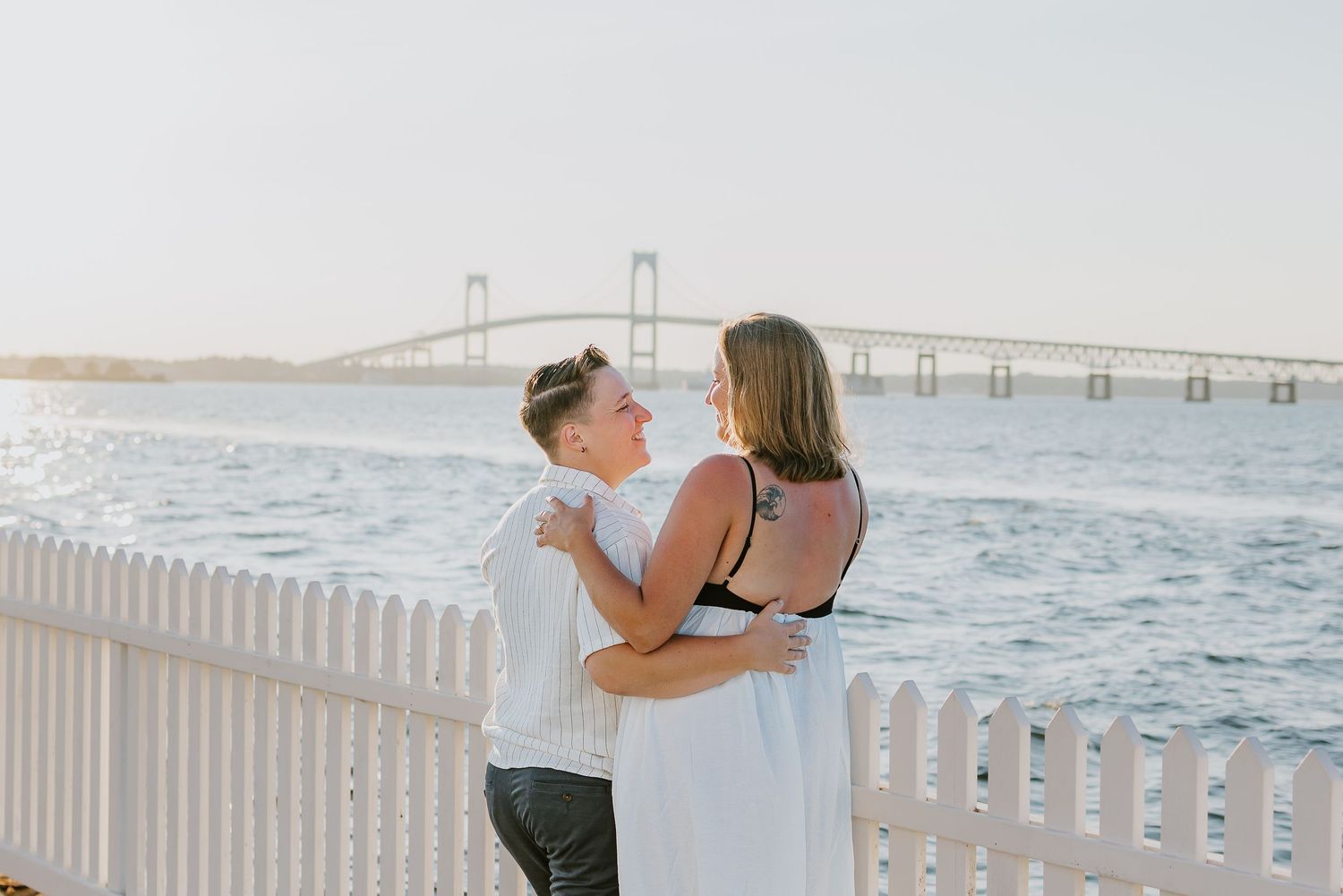 A loving LGBTQ couple embraces by a white railing overlooking the Newport Bridge and Narragansett Bay at sunset in Rhode Island
