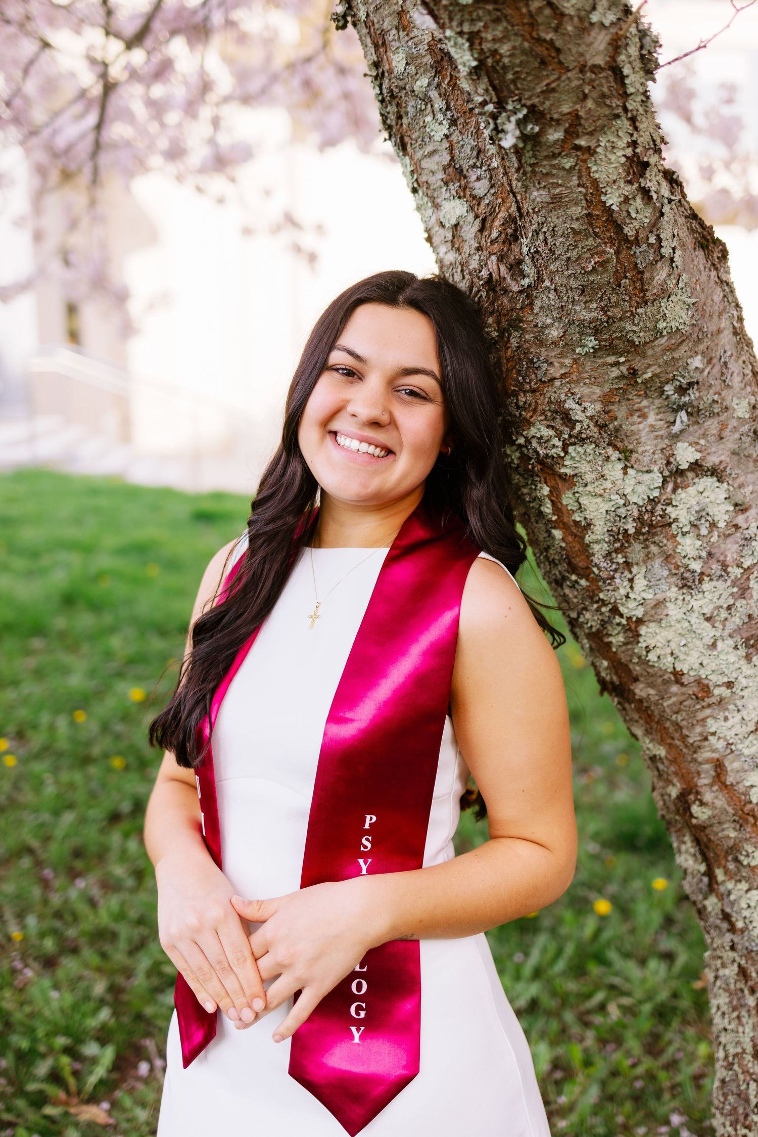 A graduate in a white dress wearing a red satin sash poses by a cherry blossom tree on a spring day at UMASS Dartmouth Massachusetts