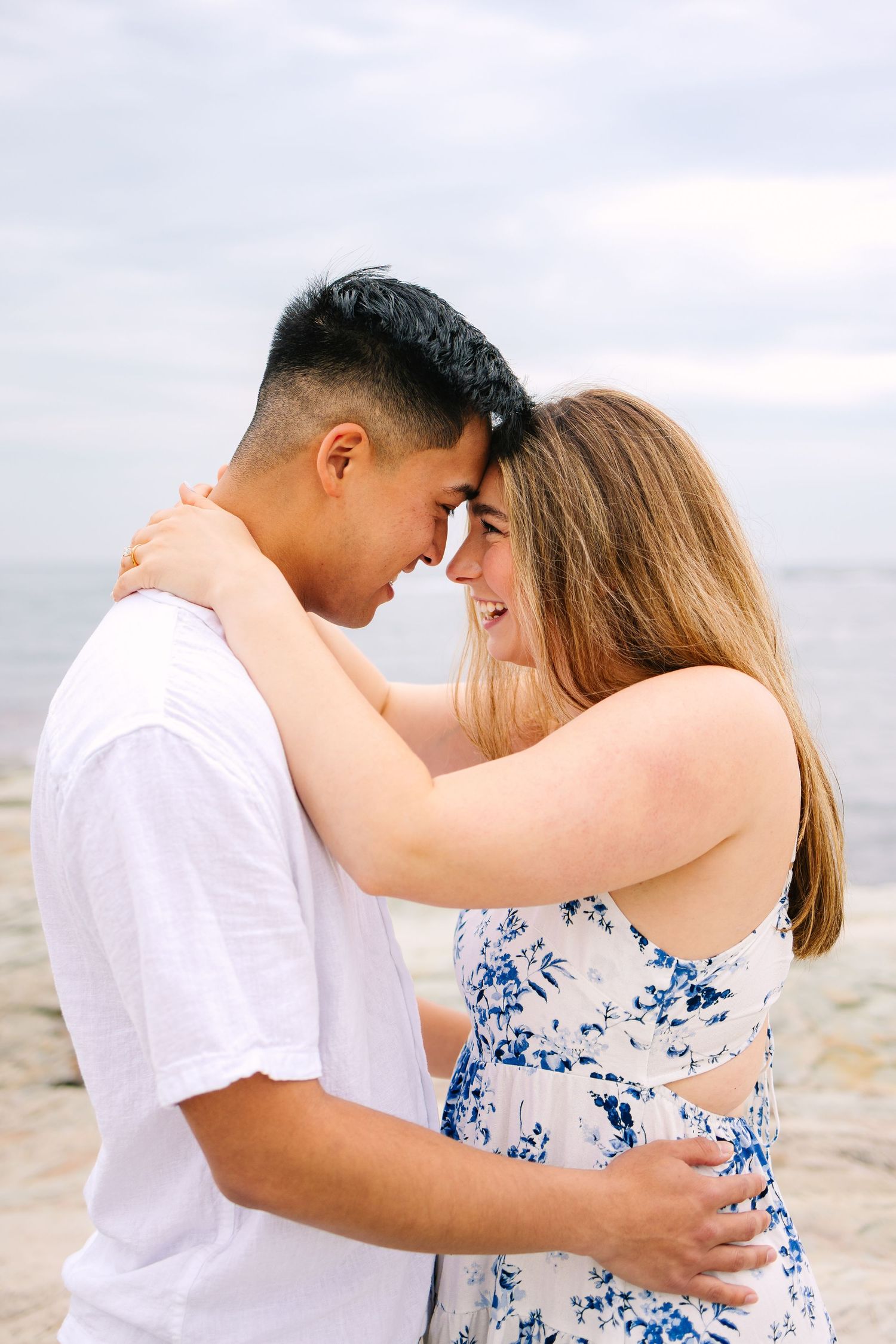 A couple embraces on a beach, touching foreheads and smiling, wearing casual summer outfits with the ocean in the background in Newport RI