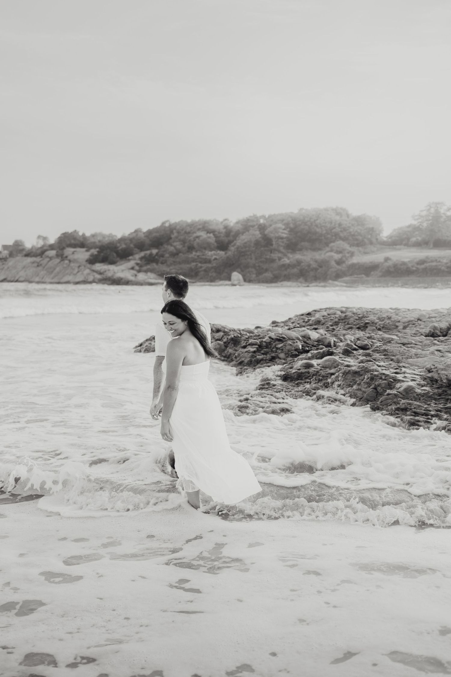 A figure in a white flowing dress walks gracefully across a rocky beach shoreline in black and white.